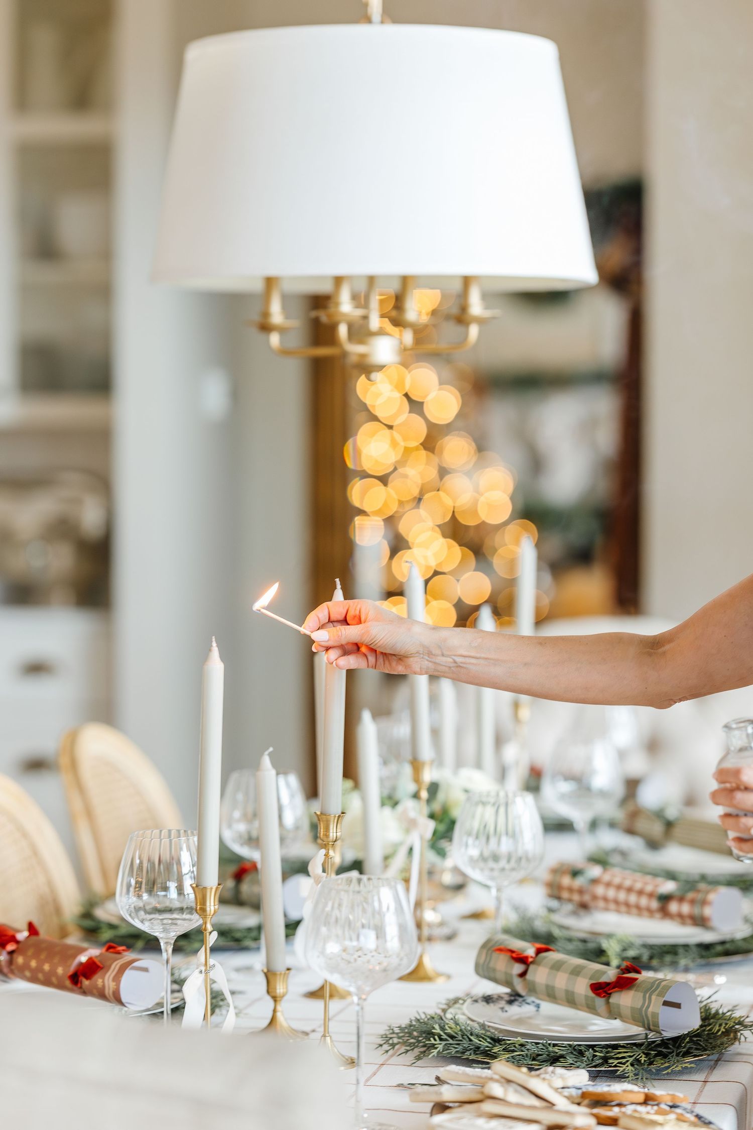 Elegant holiday table setting with white candles, wine glasses, and evergreen branches against warm bokeh lights.