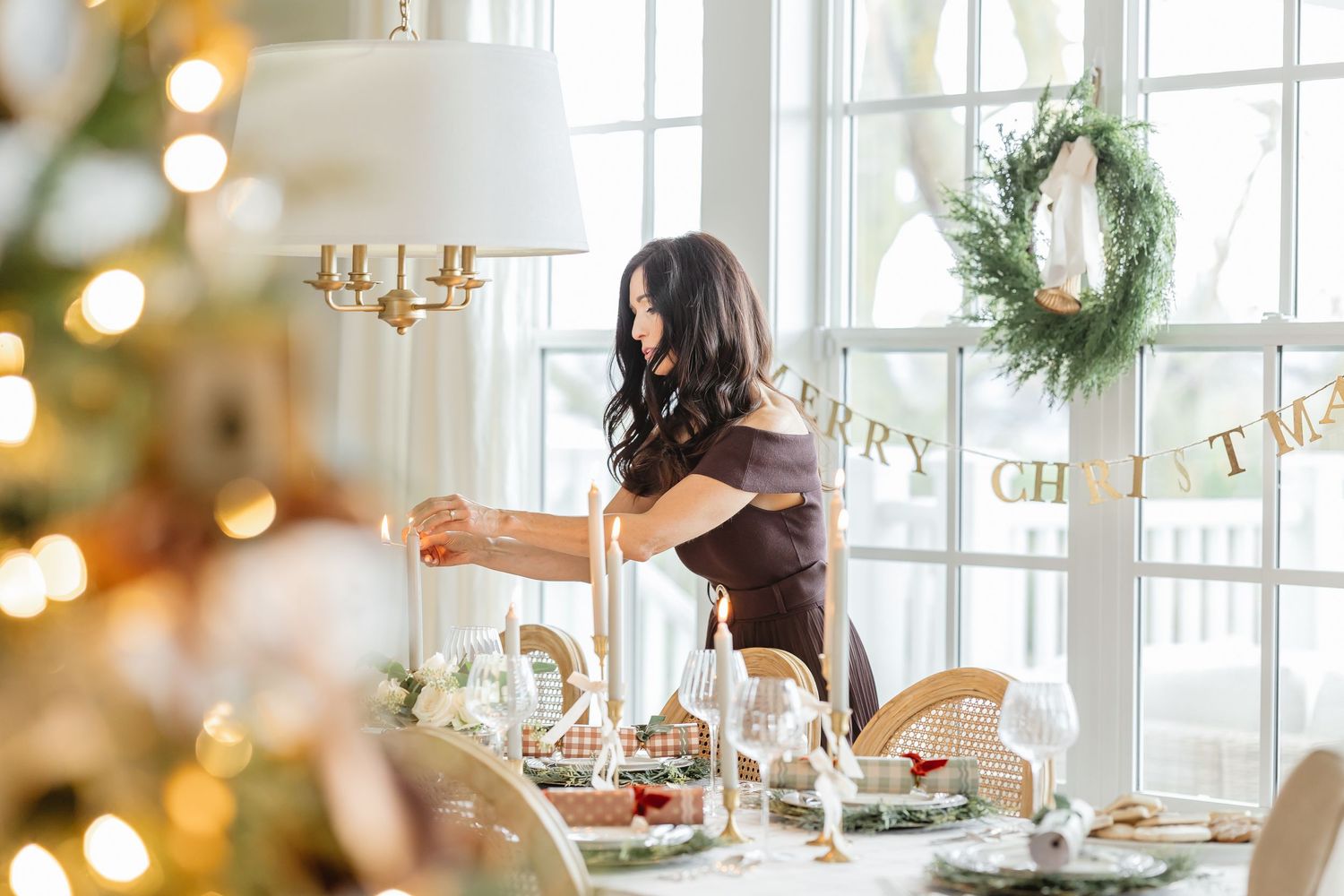 A festive dining table being set with candles and holiday decorations near a Christmas tree and window wreath.