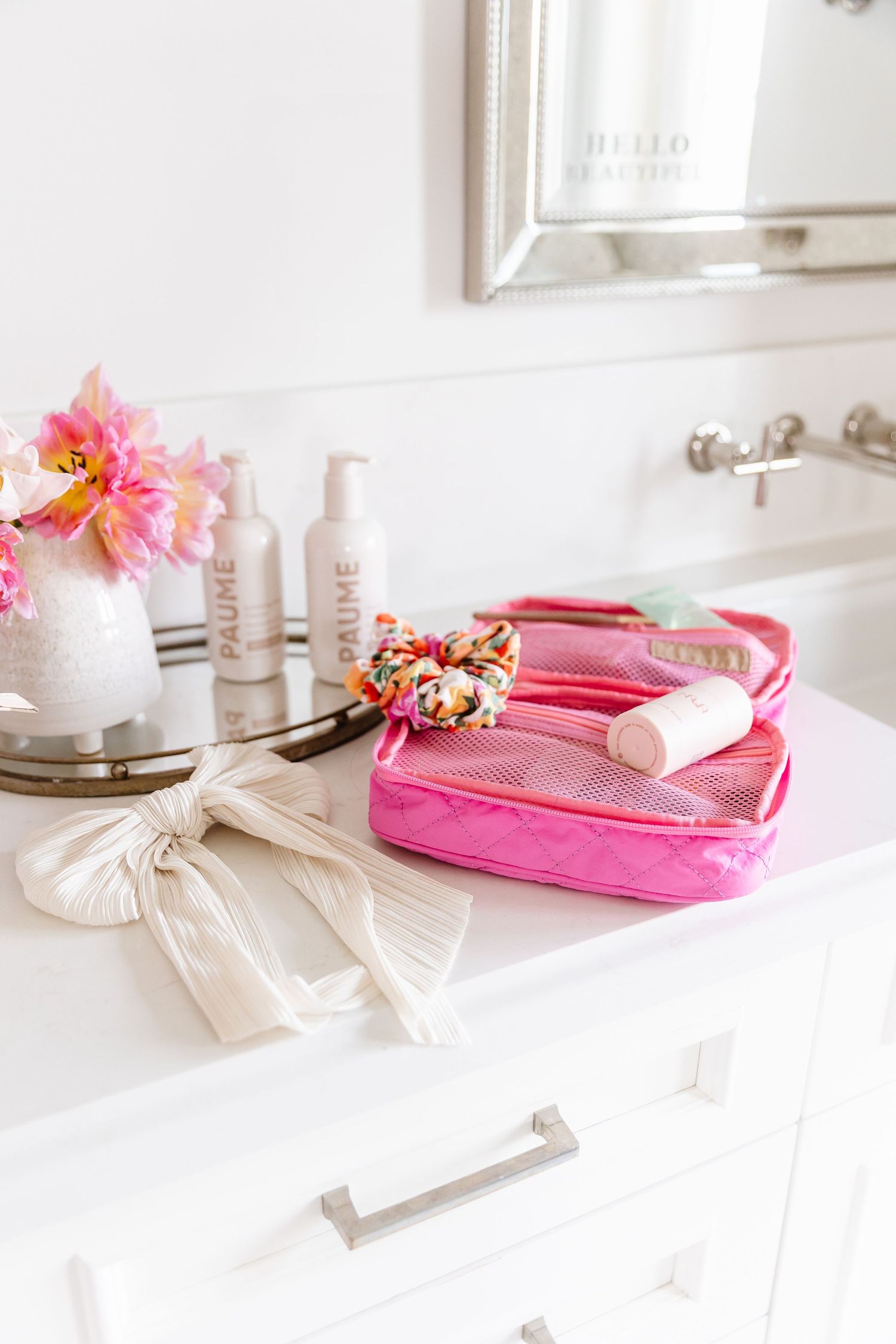 Pink velvet cosmetic bag with flowers and beauty products arranged on white vanity counter in bright natural light.