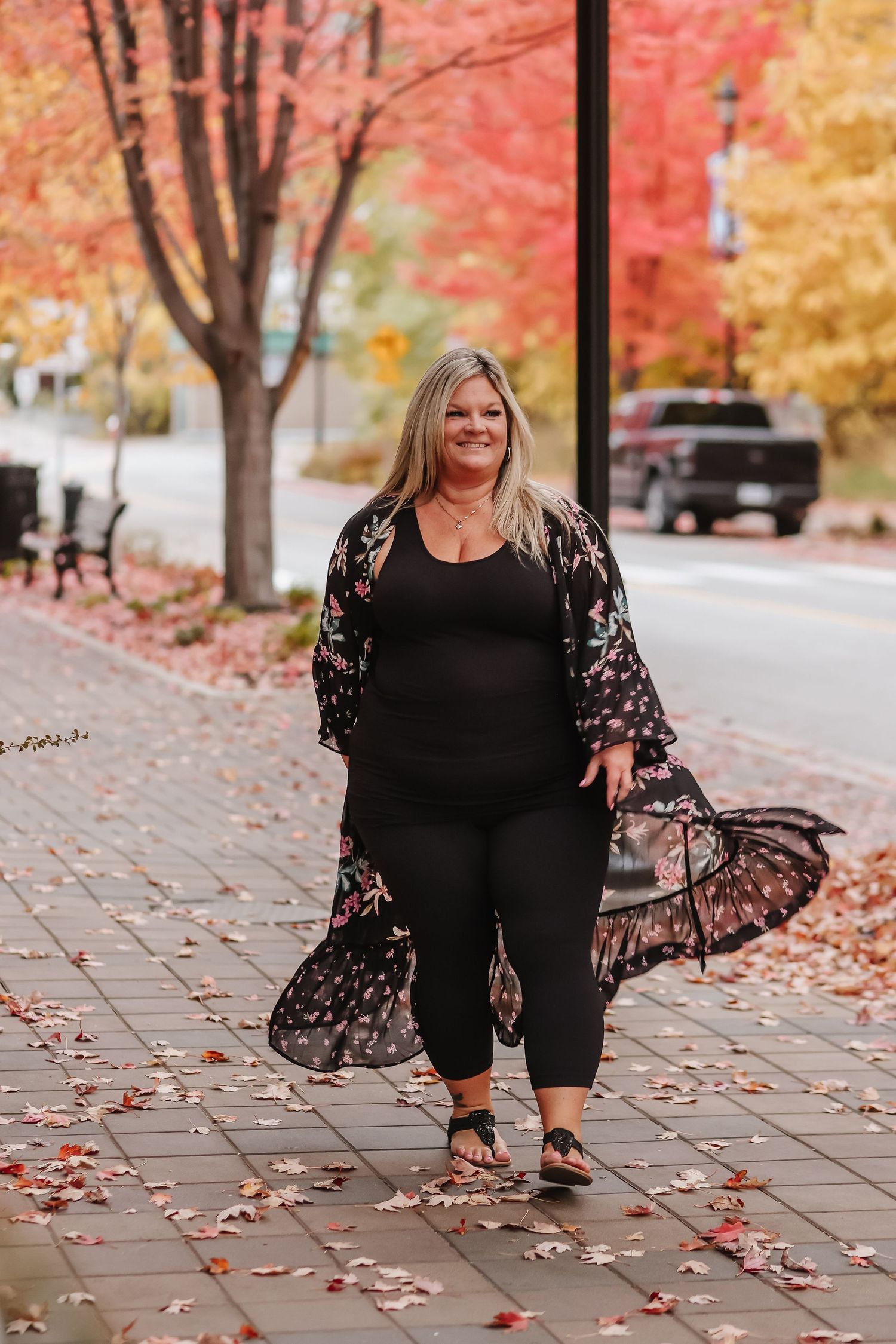 A woman in a black floral kimono and pants poses on a sidewalk with vibrant autumn foliage in the background.