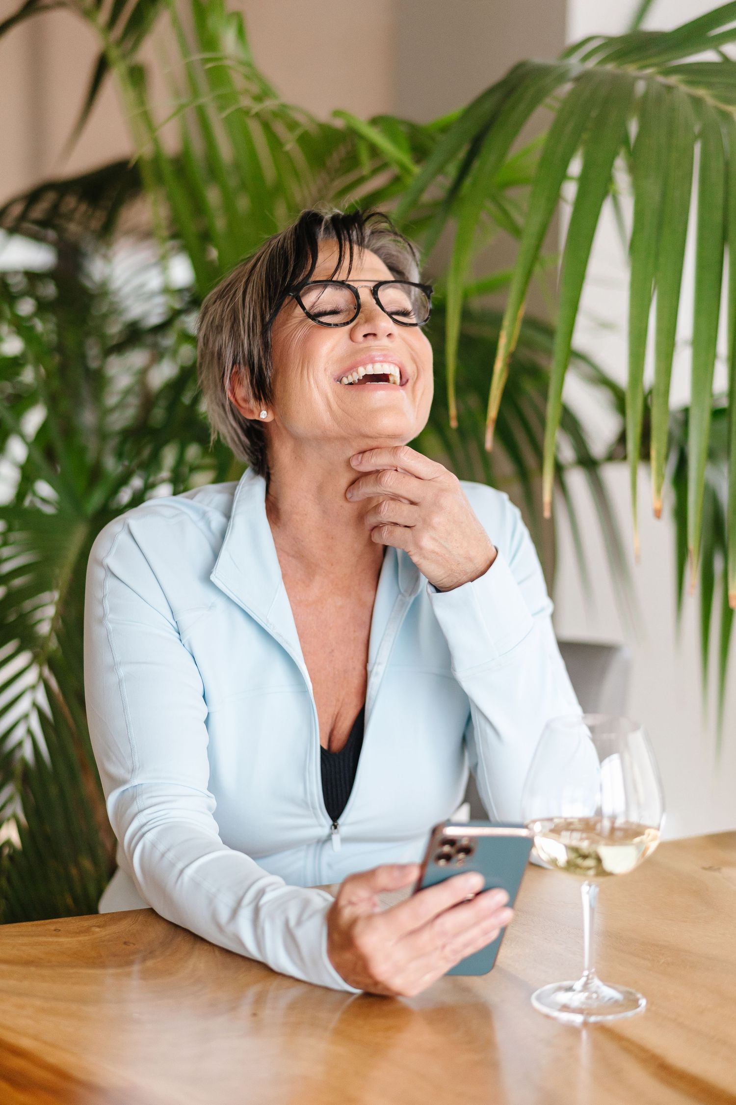 Person in white blazer and glasses laughs while looking at phone at wooden table with tropical plants in background.