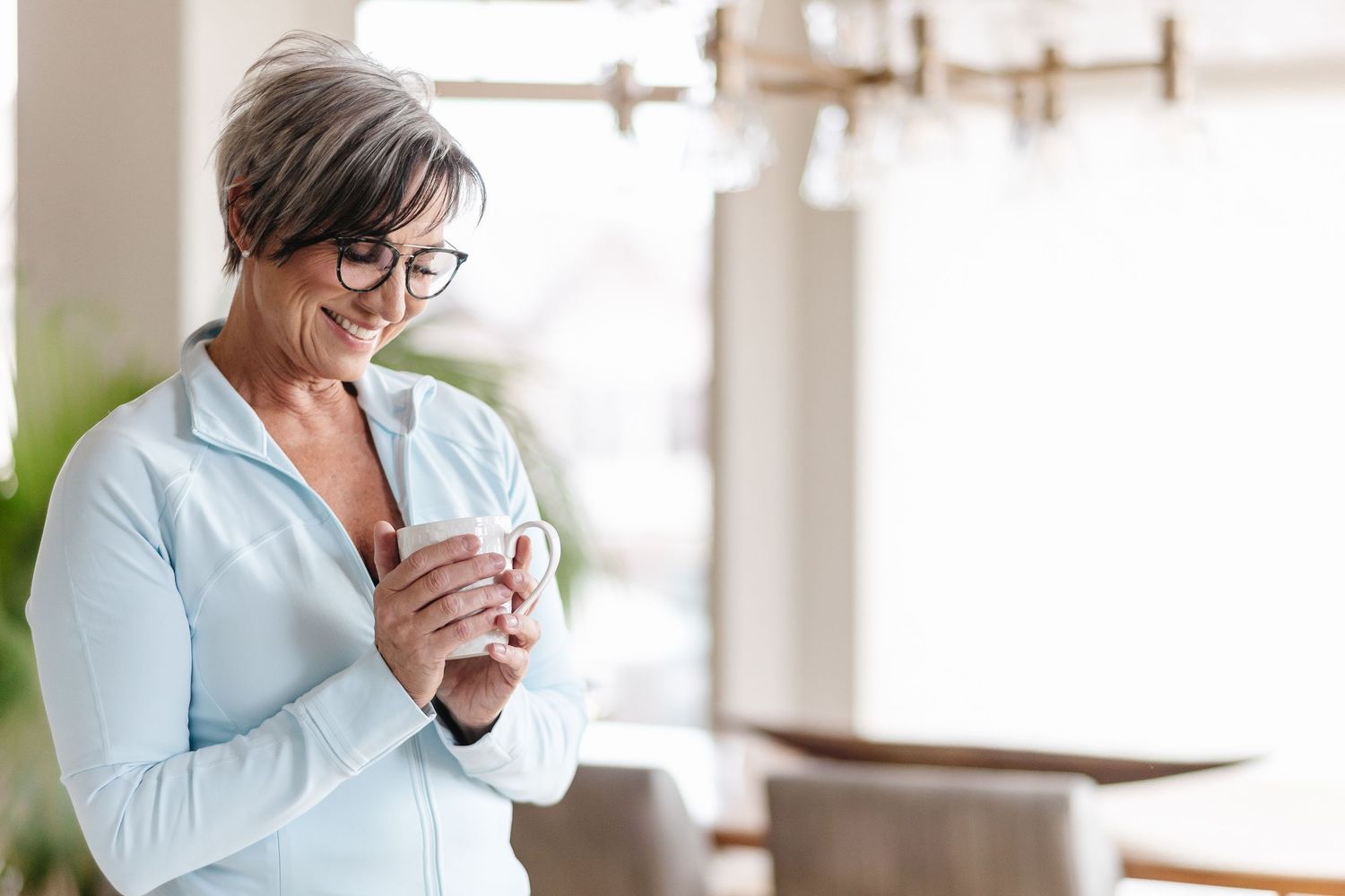 Person in light blue sweater sitting by window enjoying coffee and using smartphone in bright modern space.