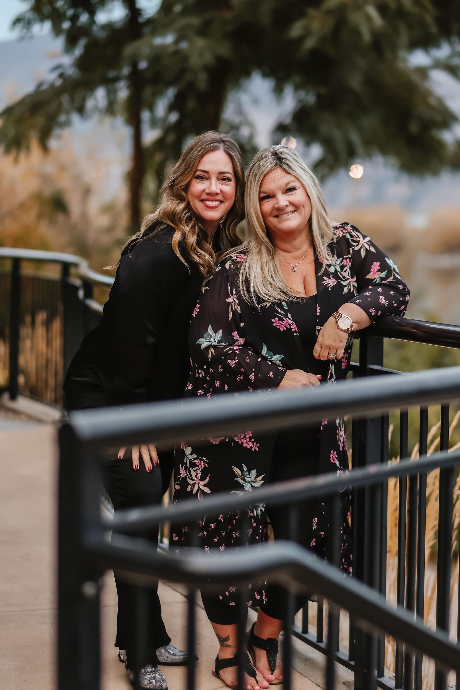 Two women pose together on a railing outdoors wearing black outfits.