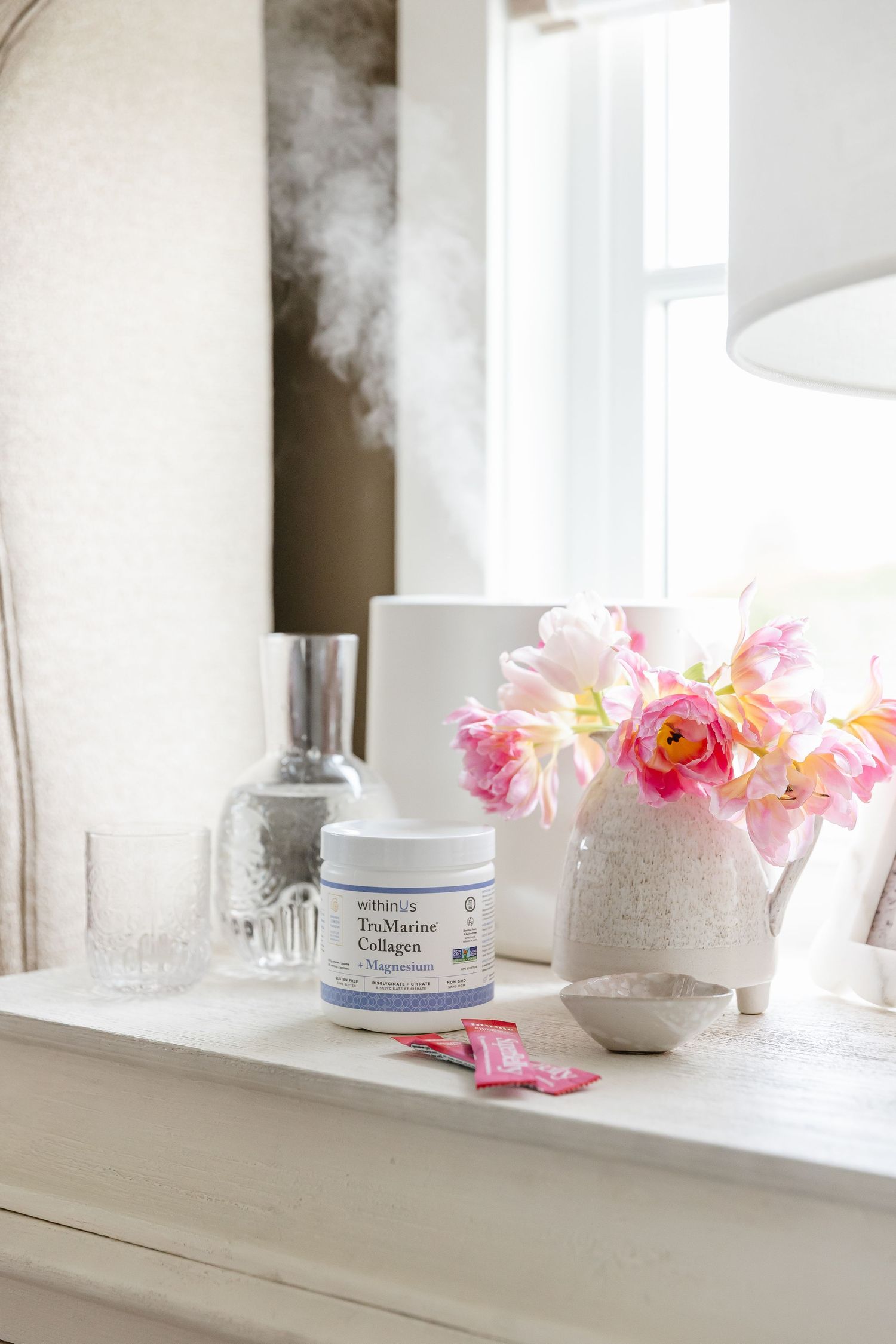 Minimalist white bathroom counter with pink flowers, skincare products, and natural light streaming through window.