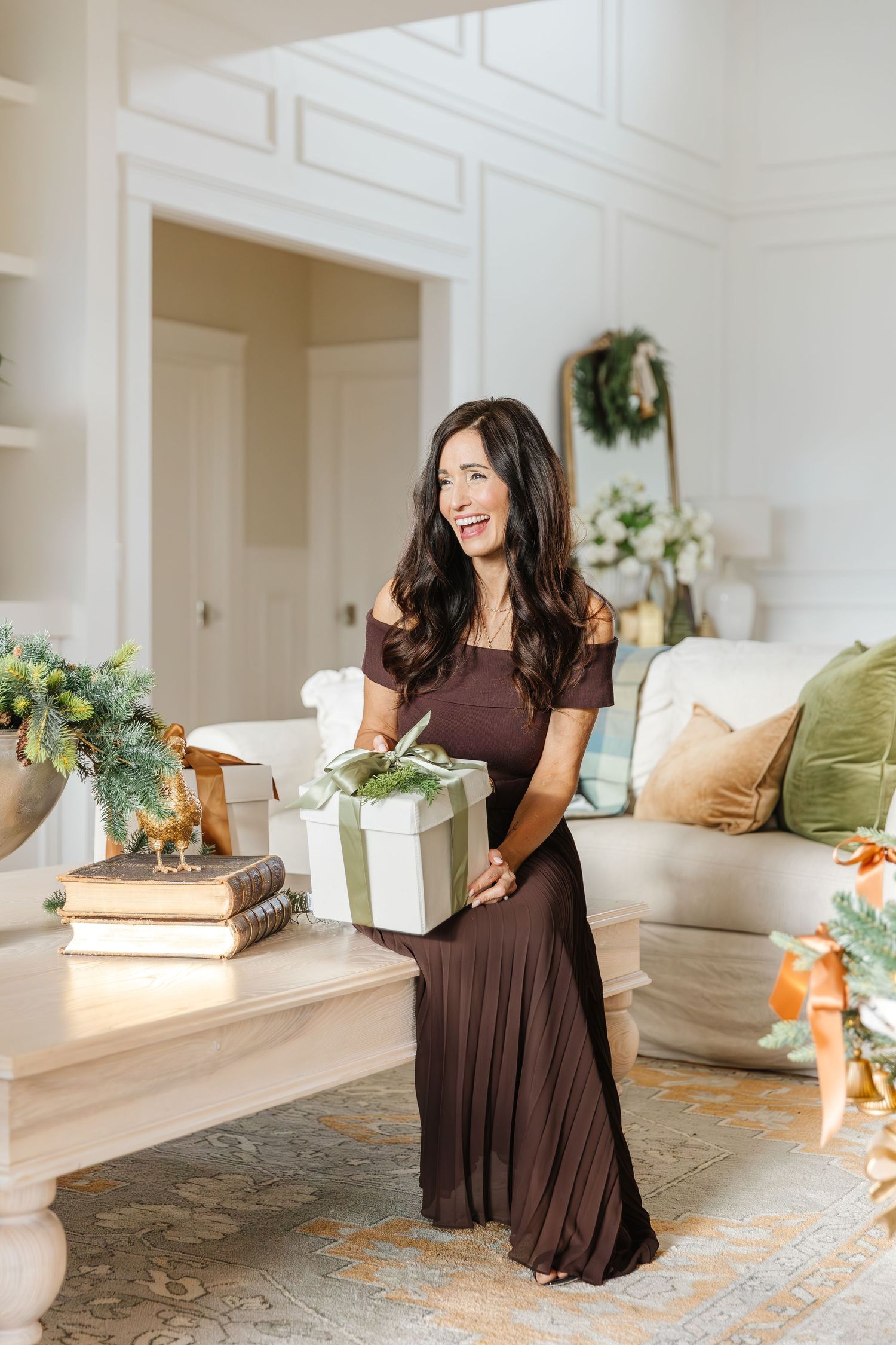 Woman in brown maxi dress holding a wrapped gift box in a festive holiday decorated living room.