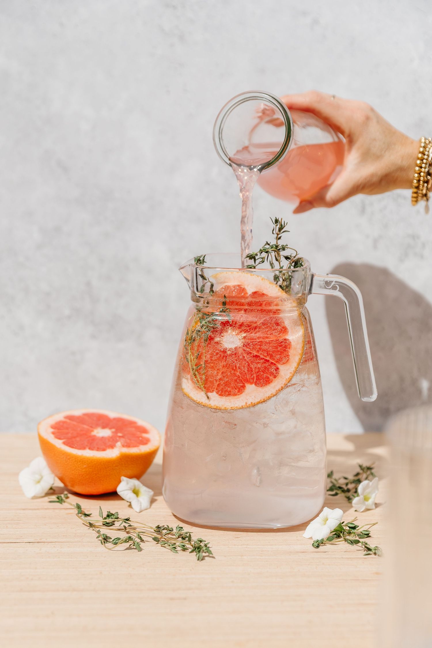 Hand pouring pink grapefruit infused water into a glass pitcher with ice, thyme, and fresh grapefruit slices.