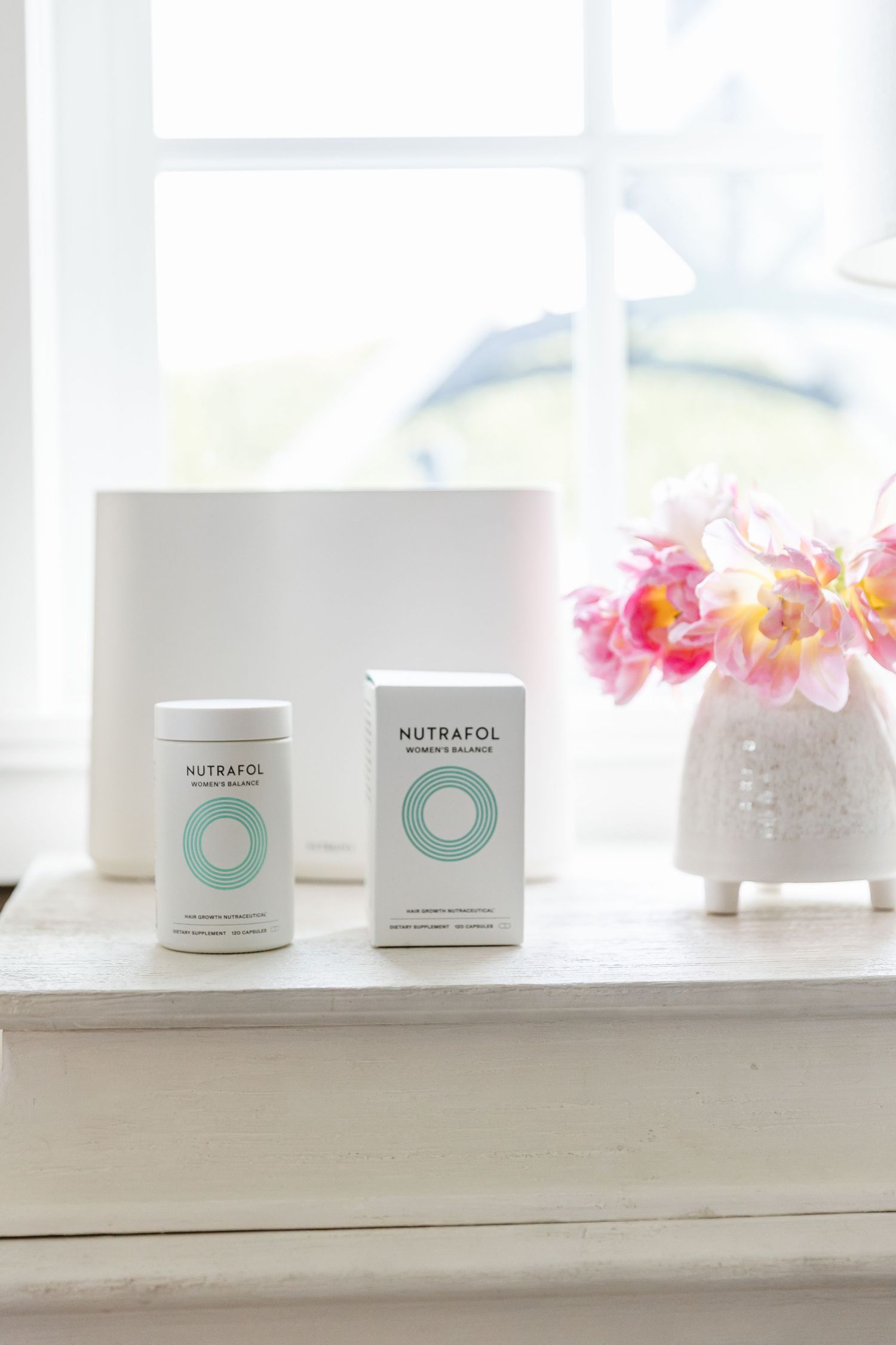 White Nutraten product containers displayed on windowsill with pink flowers in decorative vase creating serene composition.