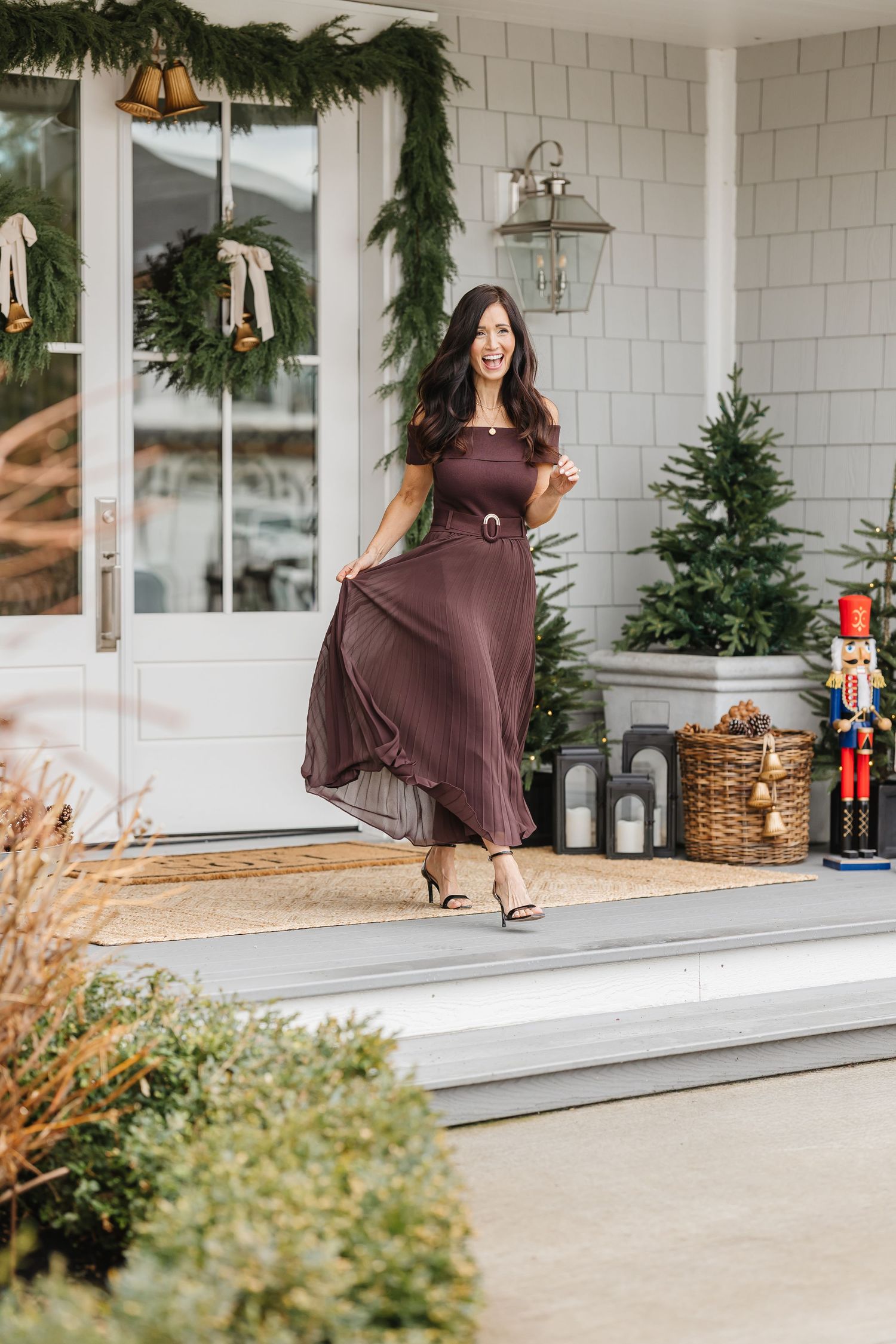 A festive holiday porch decorated with garland and a nutcracker features a flowing brown dress against white shingle siding.