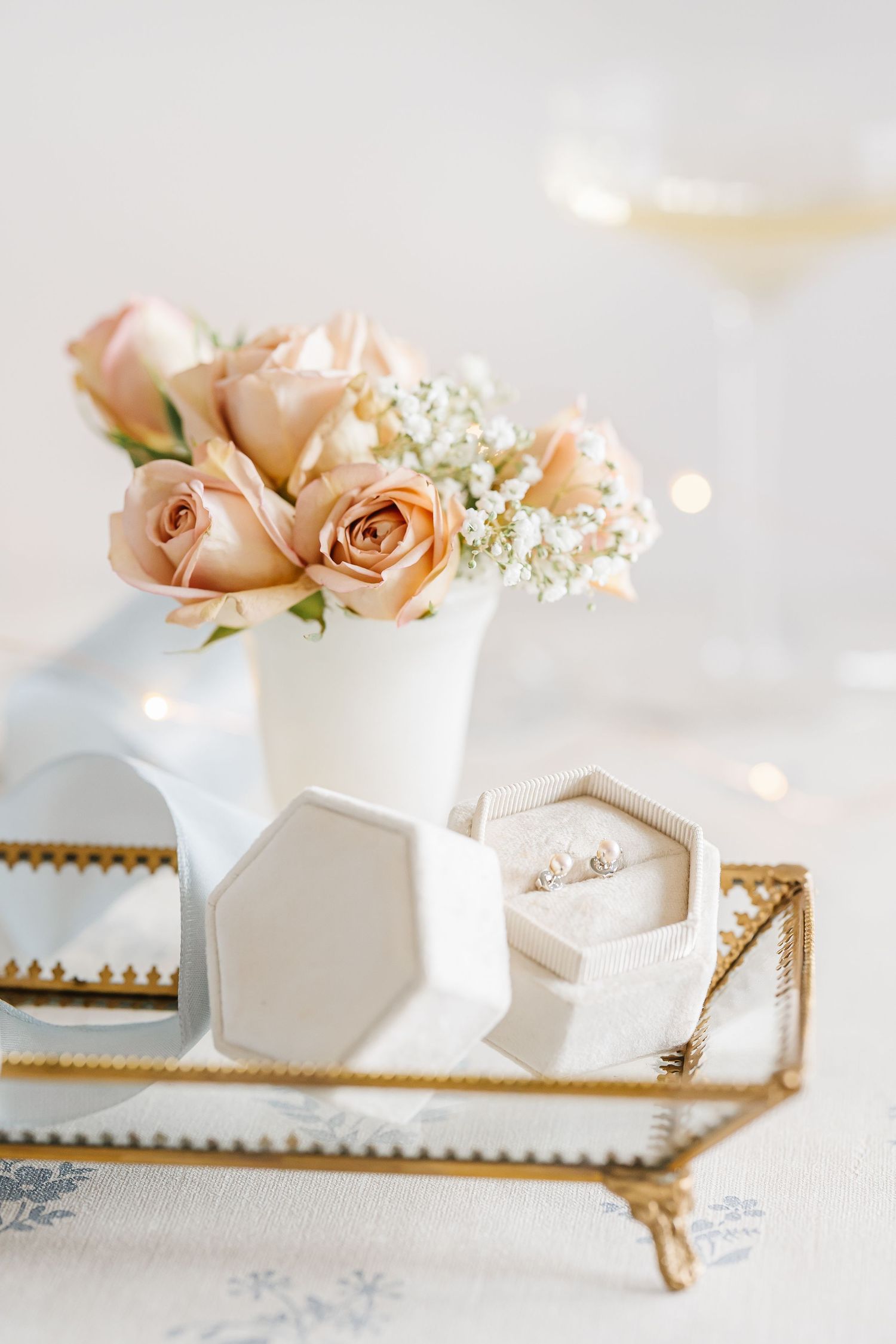 Blush roses in white vase on gold mirrored tray with hexagon ring box and wine glass.
