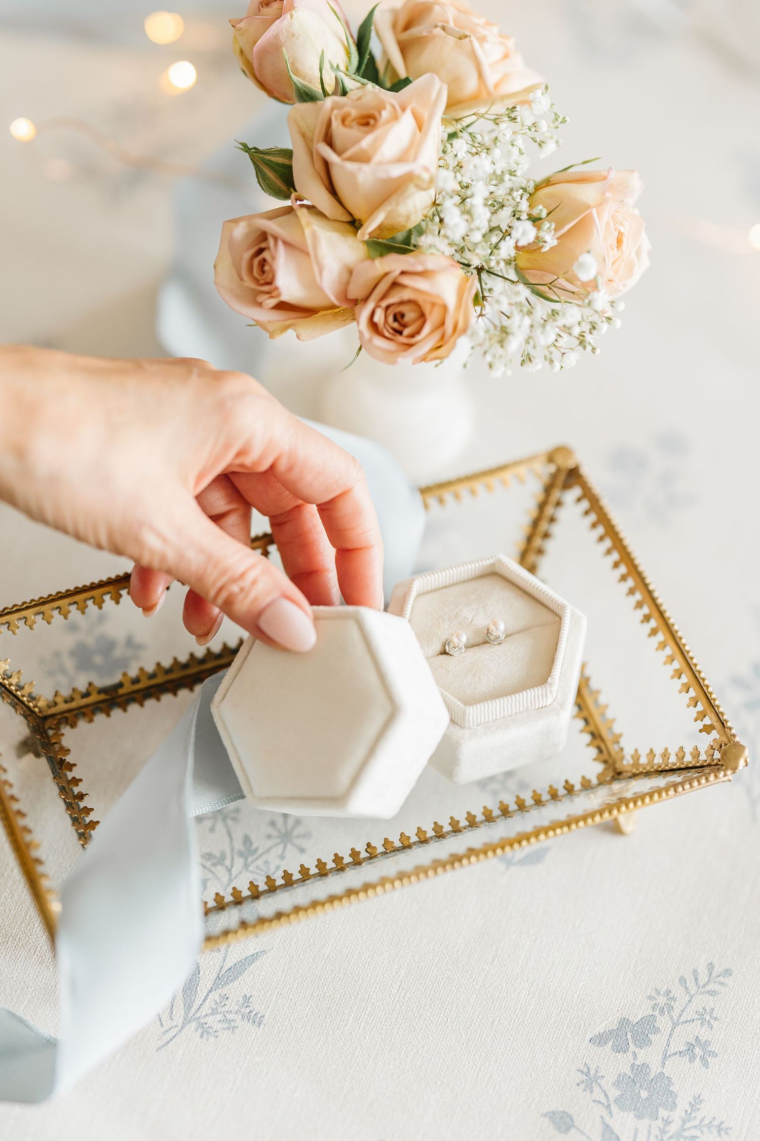Hand opening a white hexagonal ring box with diamond ring on gold tray beside peach roses in white vase.