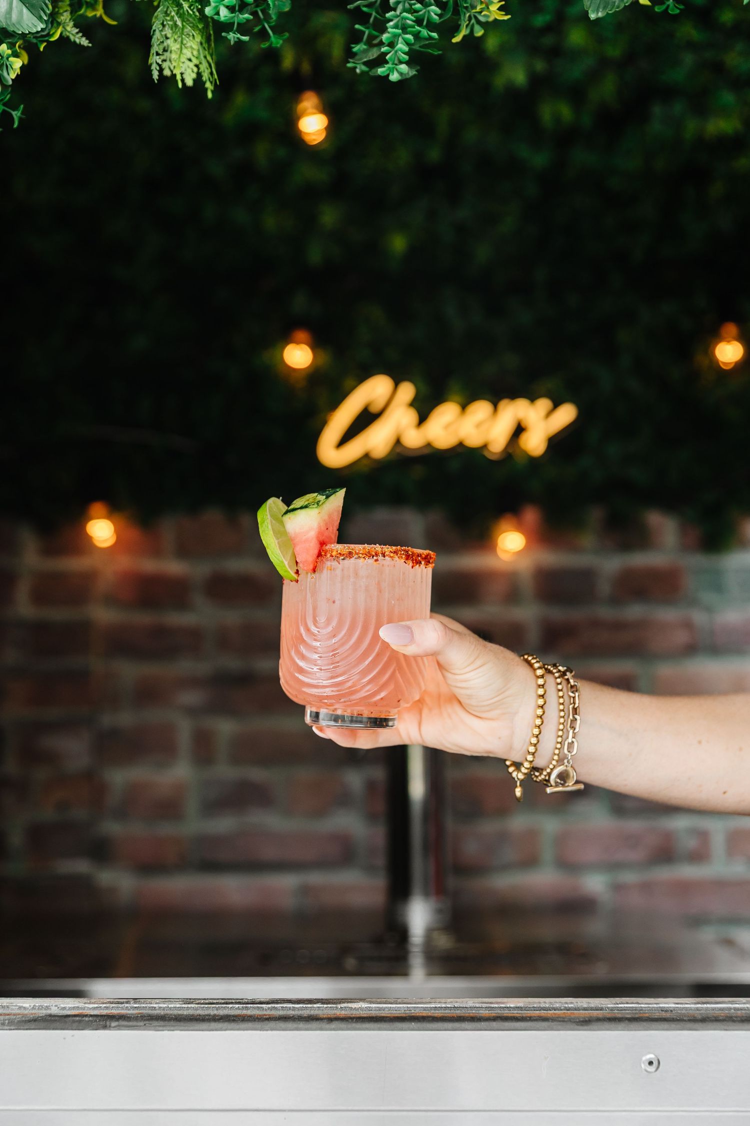 Hand holding a pink cocktail with chili rim and basil garnish, with a glowing Cheers neon sign backdrop.