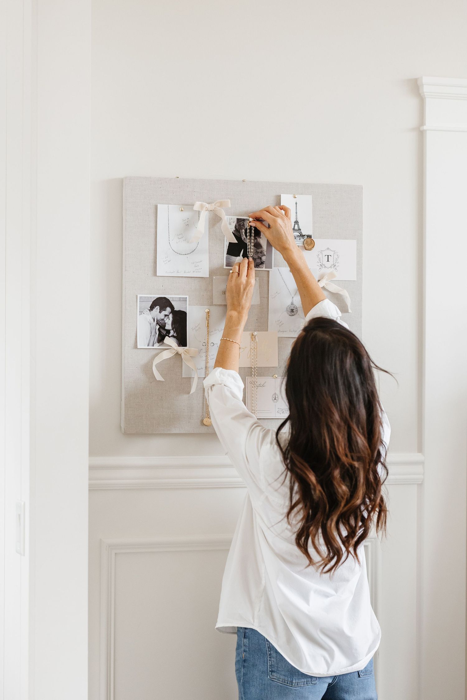 Woman with long brunette hair arranging photos and mementos on a fabric mood board mounted on a white wall.