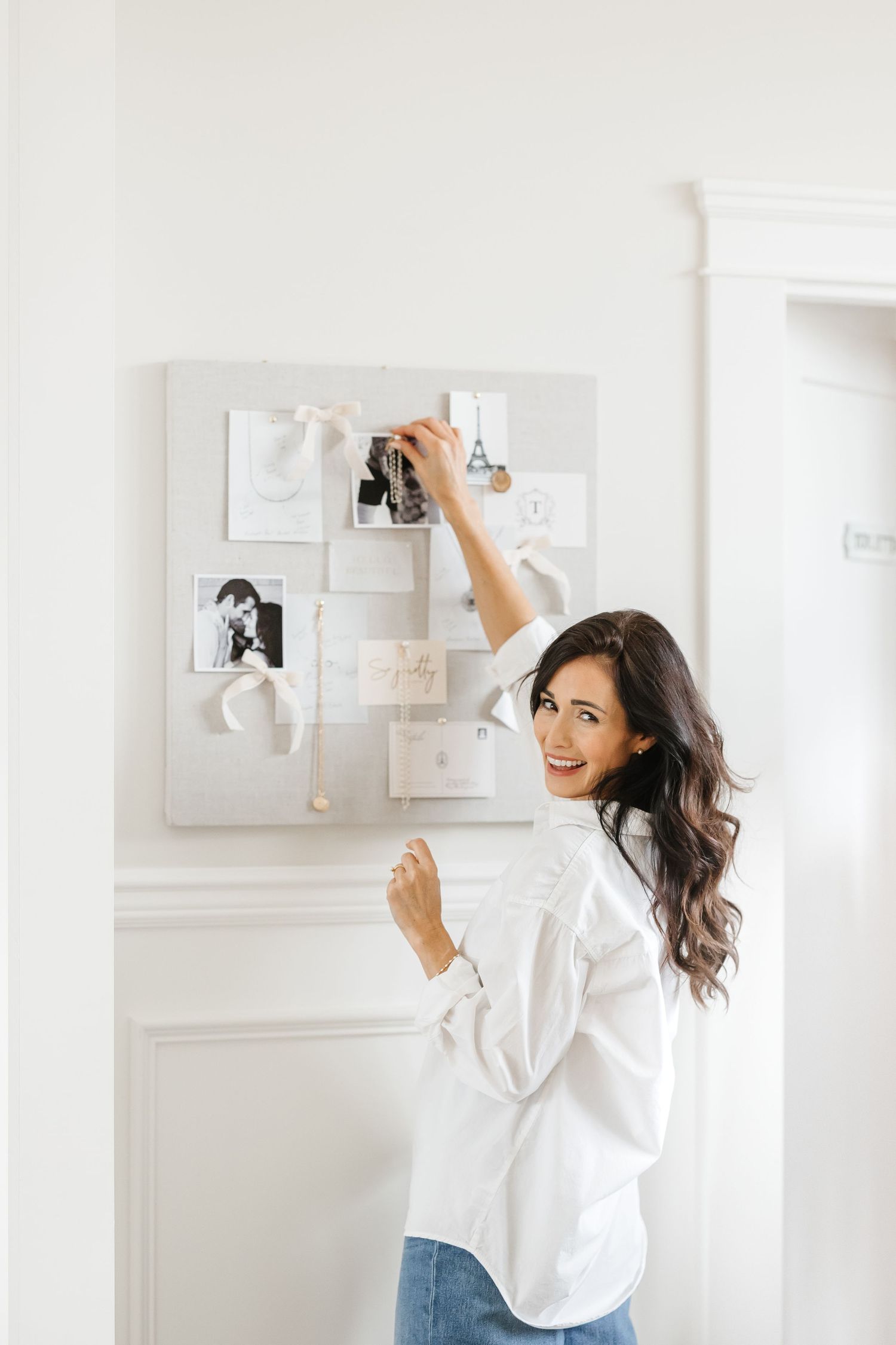 Smiling woman in white shirt pinning photos to a decorative mood board on a bright white wall.
