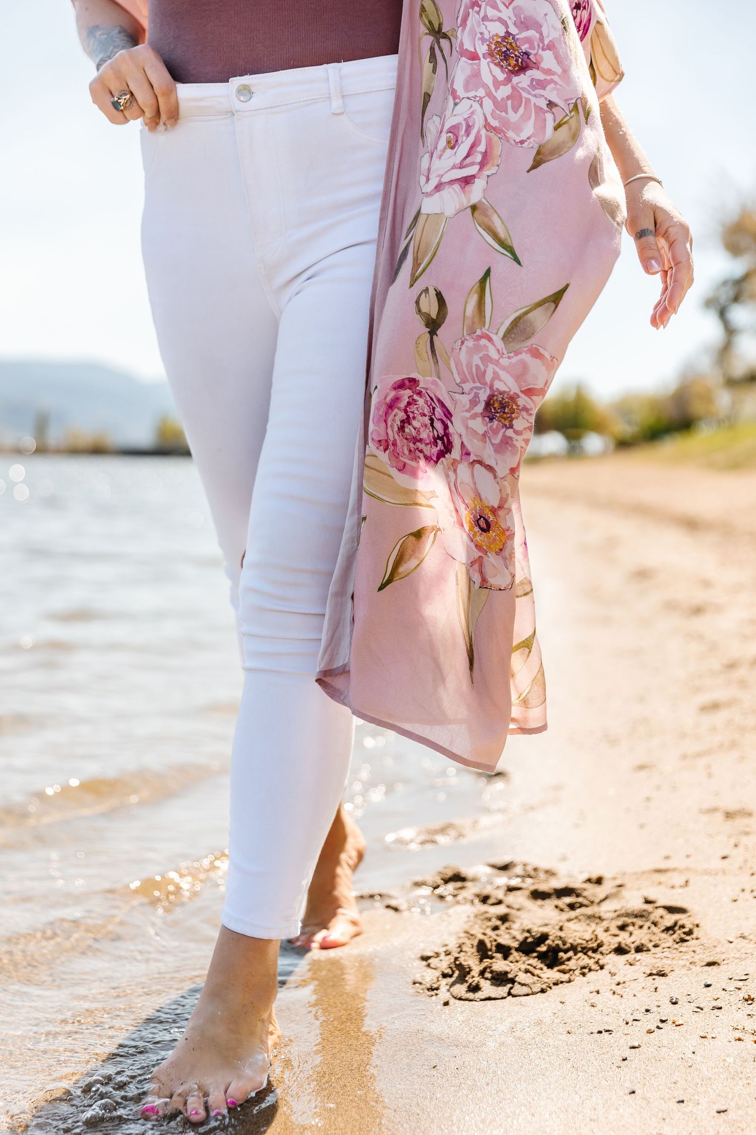 Person wearing white pants and floral top walking along a sandy beach at sunset.