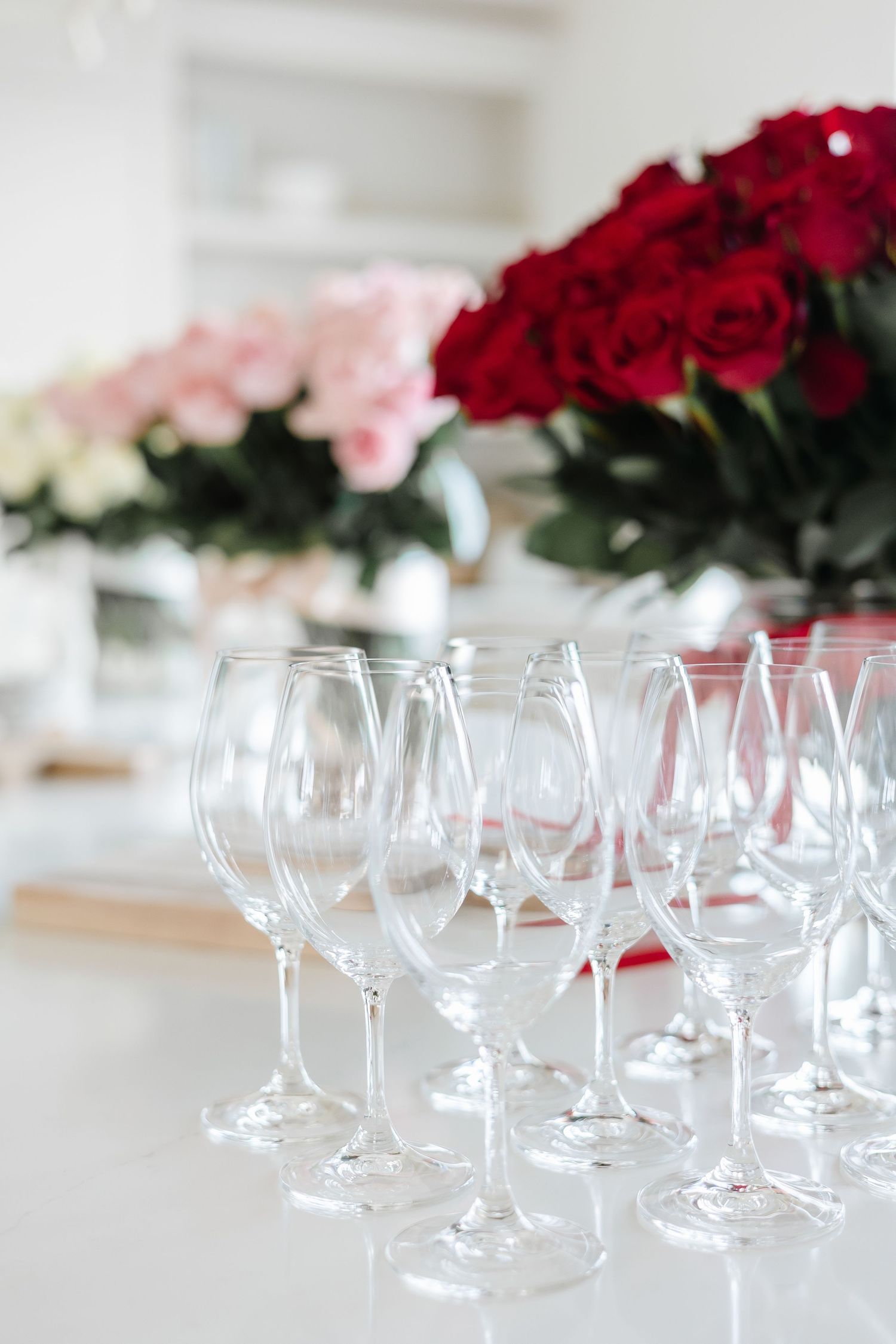 Empty crystal wine glasses arranged on a white table with bouquets of red and pink roses in the background.