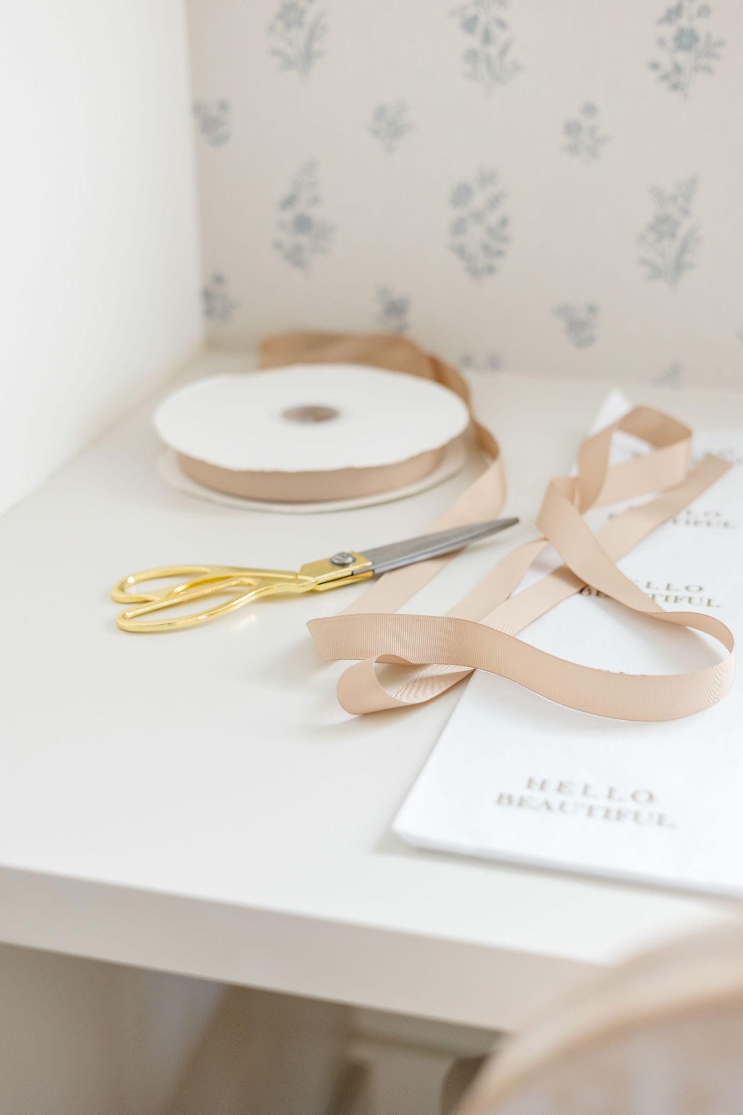 Gold scissors, blush satin ribbon on a spool, and a Hello Beautiful card arranged on a white desk.