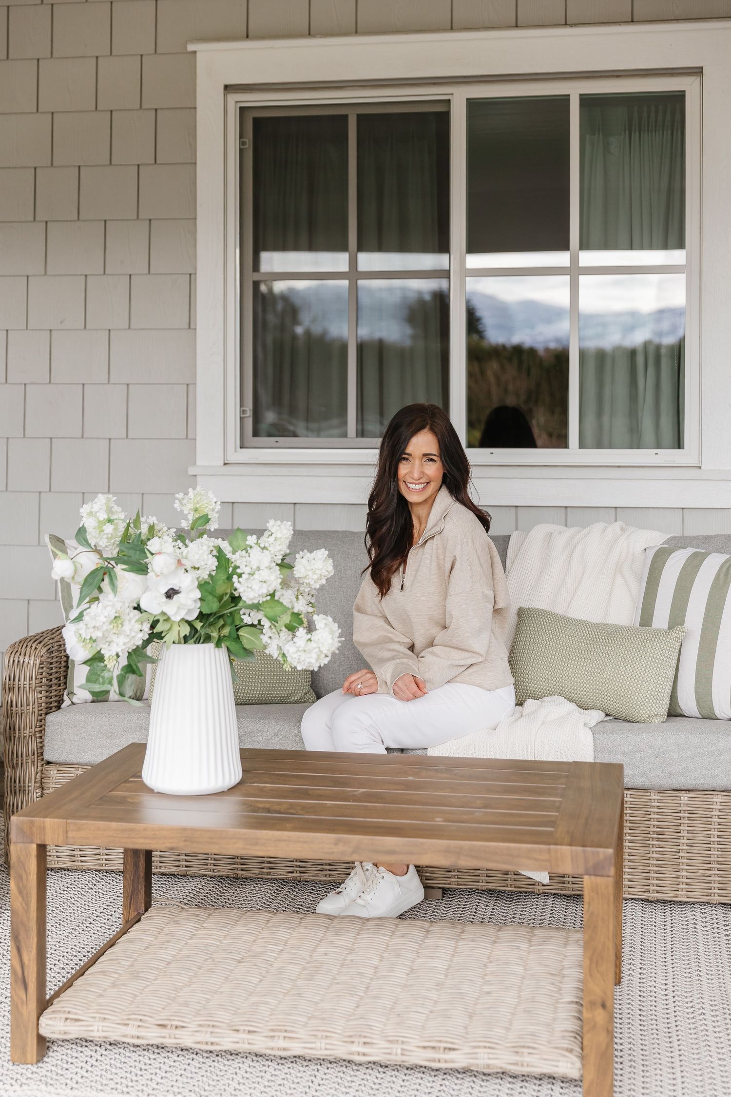 Woman sitting on outdoor wicker sofa with green striped pillows, wood coffee table, and white floral arrangement on porch.