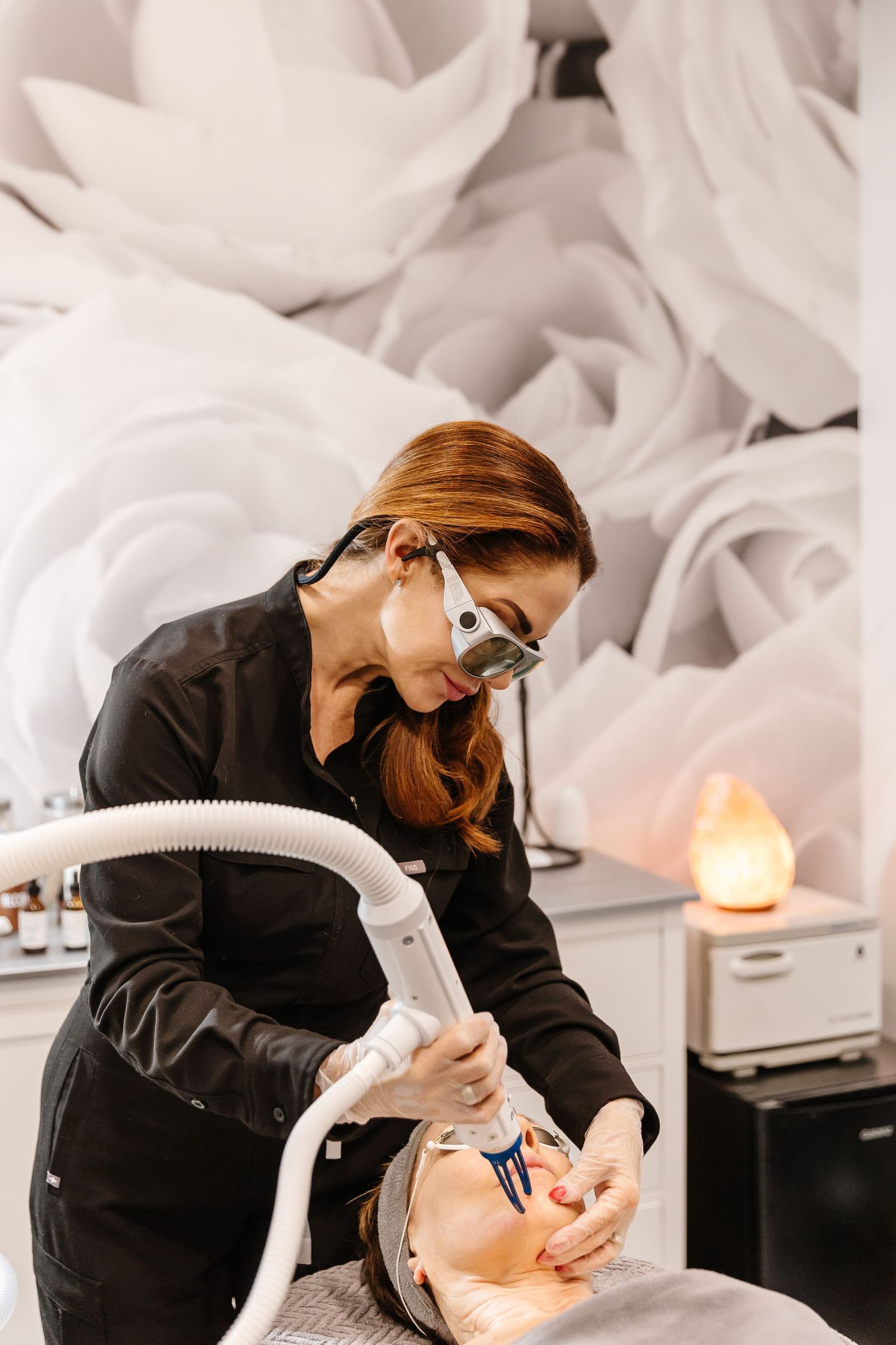 Professional esthetician performing facial treatment on client in white spa setting with candle lighting.