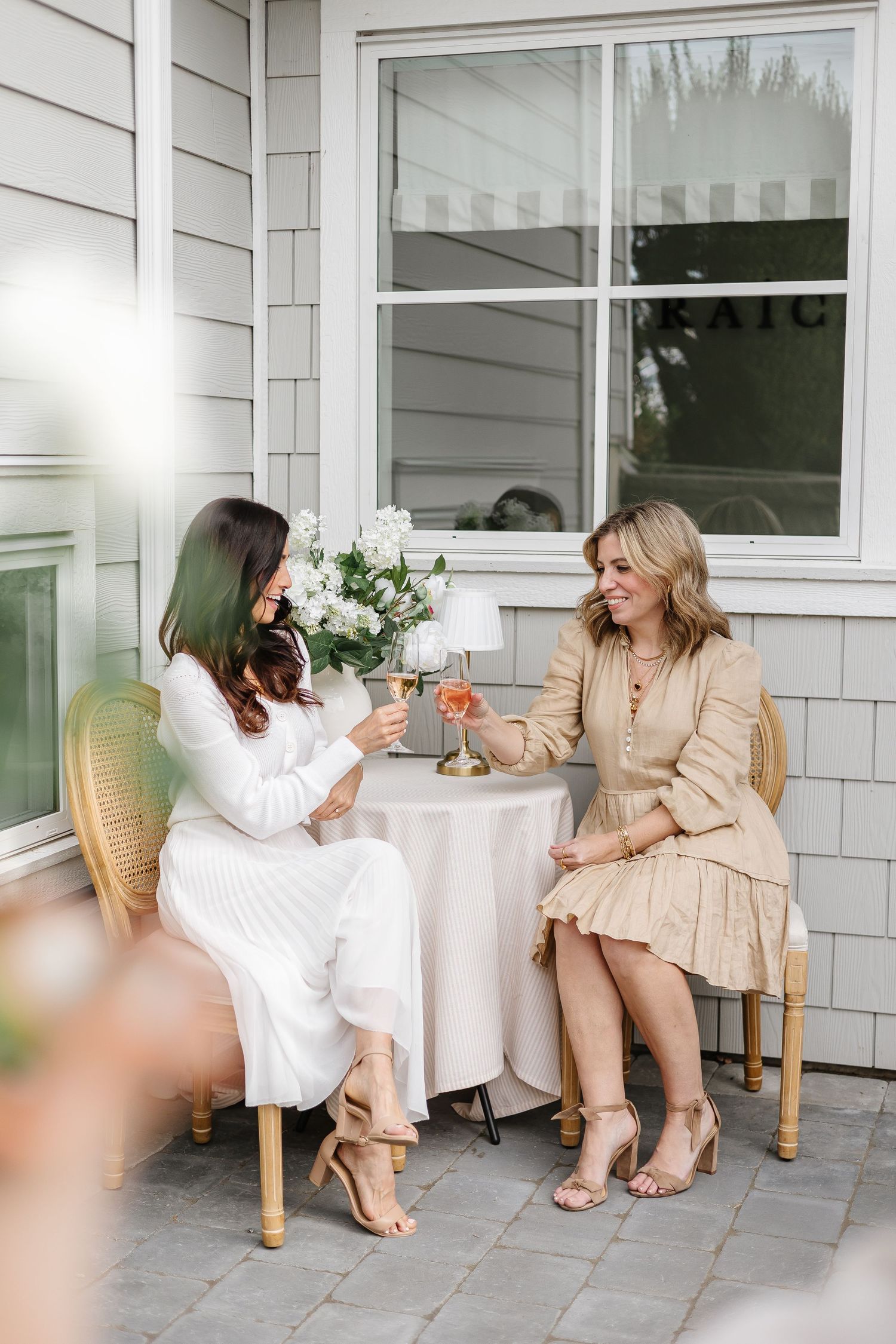 Two women toasting with rosé wine at an outdoor patio table adorned with white flowers and a small lamp.
