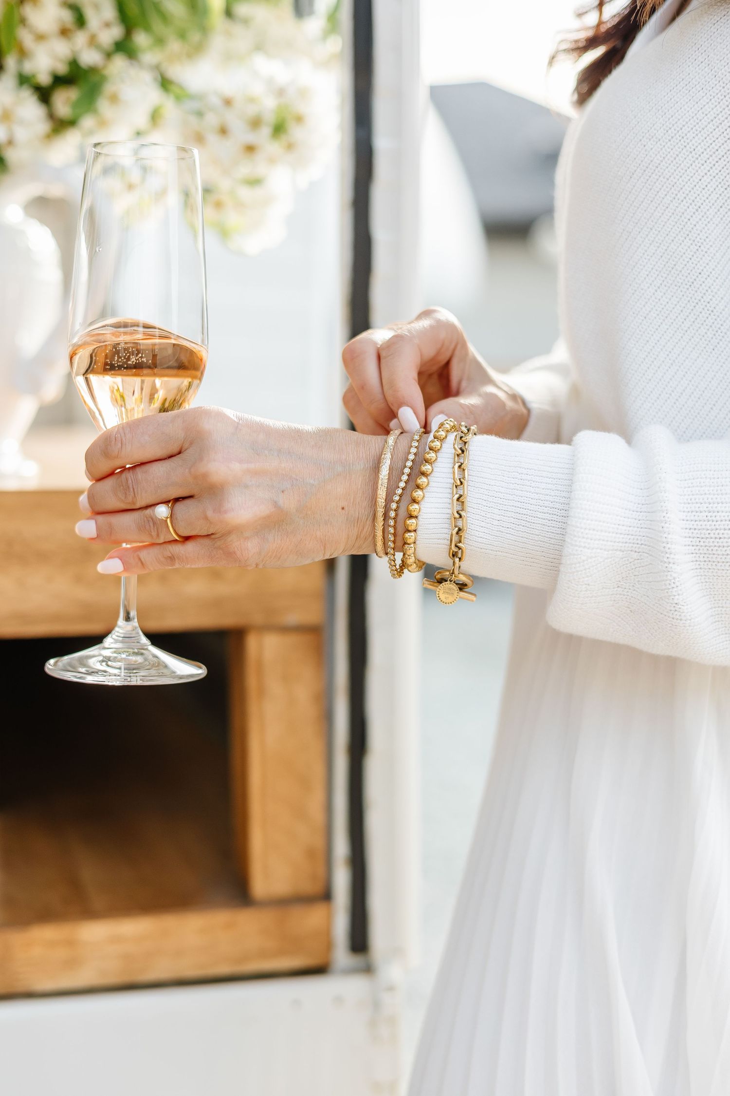 Hands wearing white sleeves hold delicate wine glasses with rose-colored beverages and gold jewelry accents.