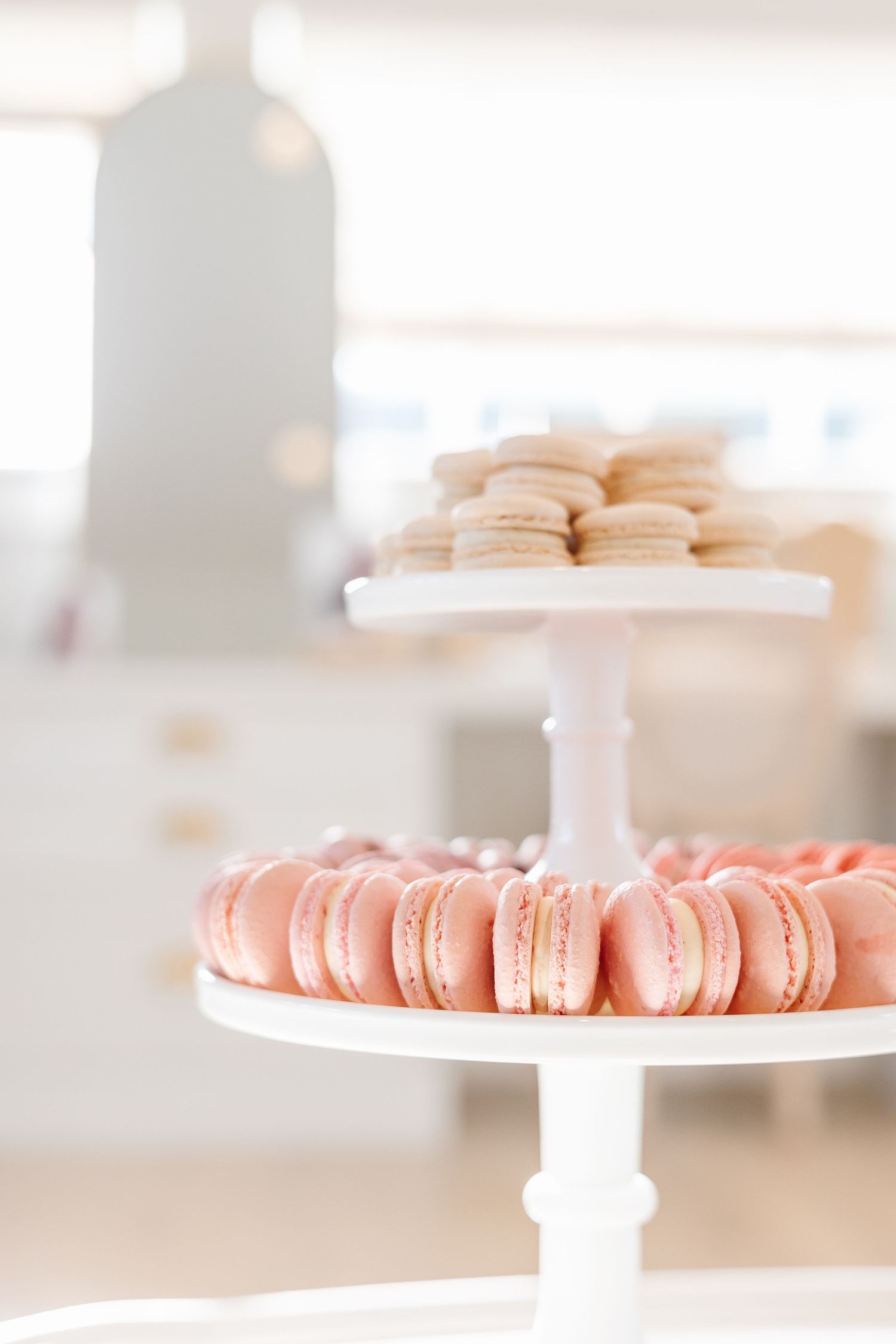 Pink and beige macarons displayed on white cake stands in a bright, airy setting.