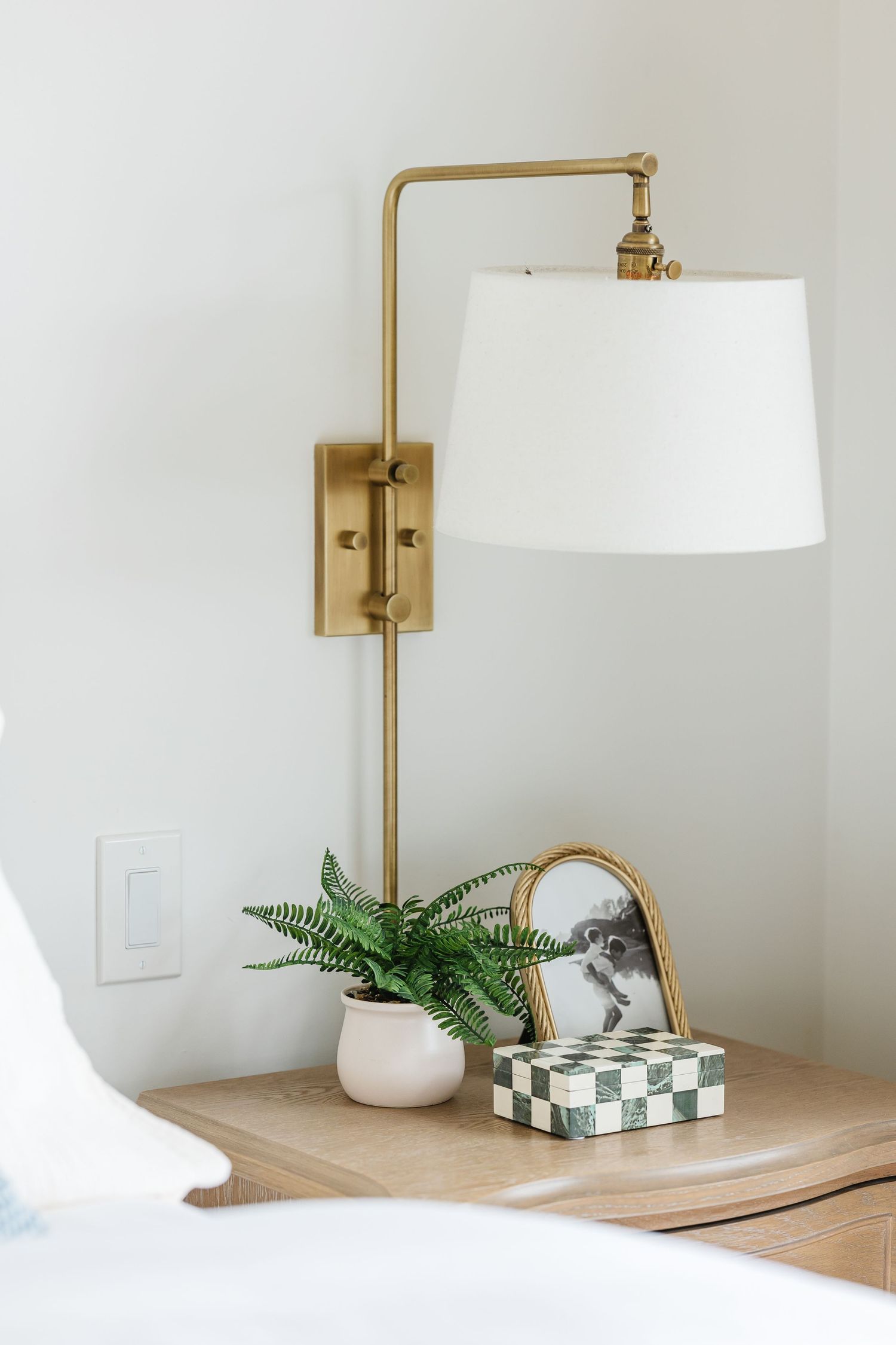 Gold brass swing arm wall sconce with white shade above wooden nightstand with fern plant and checkered box.