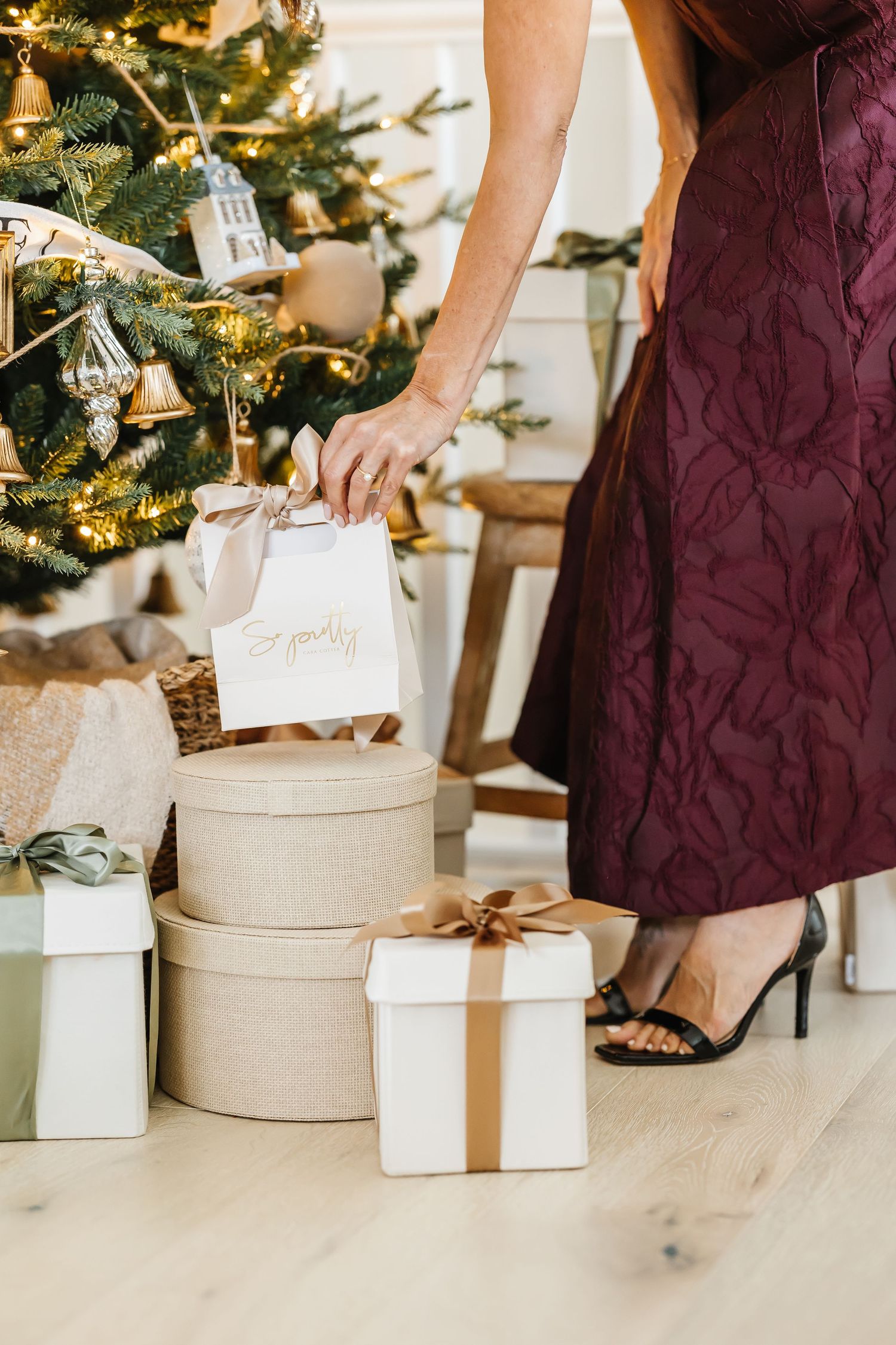 Woman in burgundy dress placing gifts under a decorated Christmas tree with gold ornaments and wrapped presents.