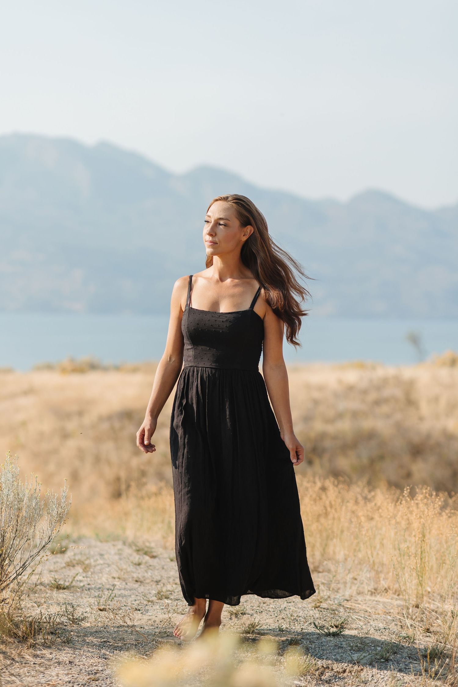 Woman in black sundress stands in golden field with mountains in background during sunset.