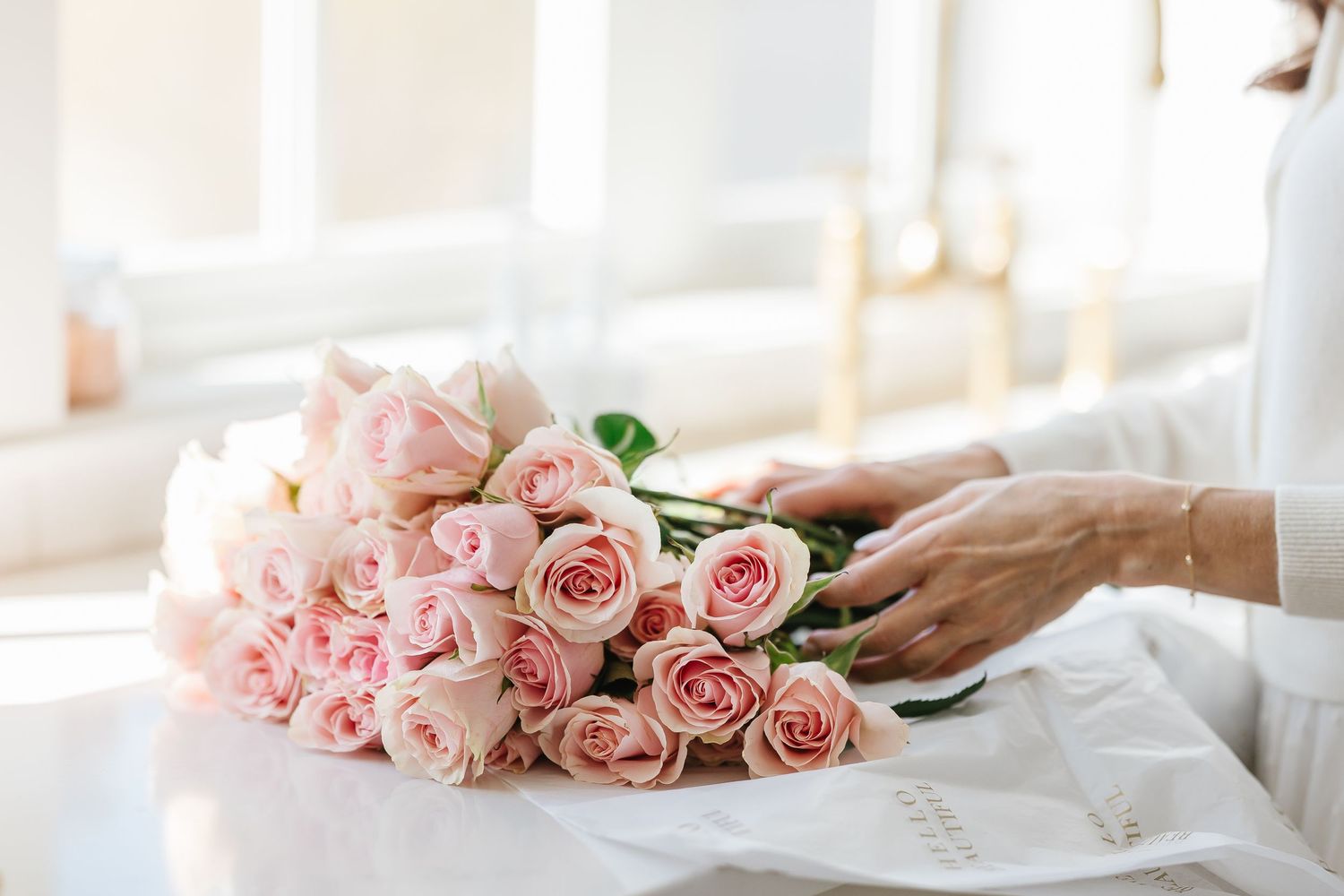 Person arranging a bouquet of soft pink roses on a white marble table in a bright, elegant room.