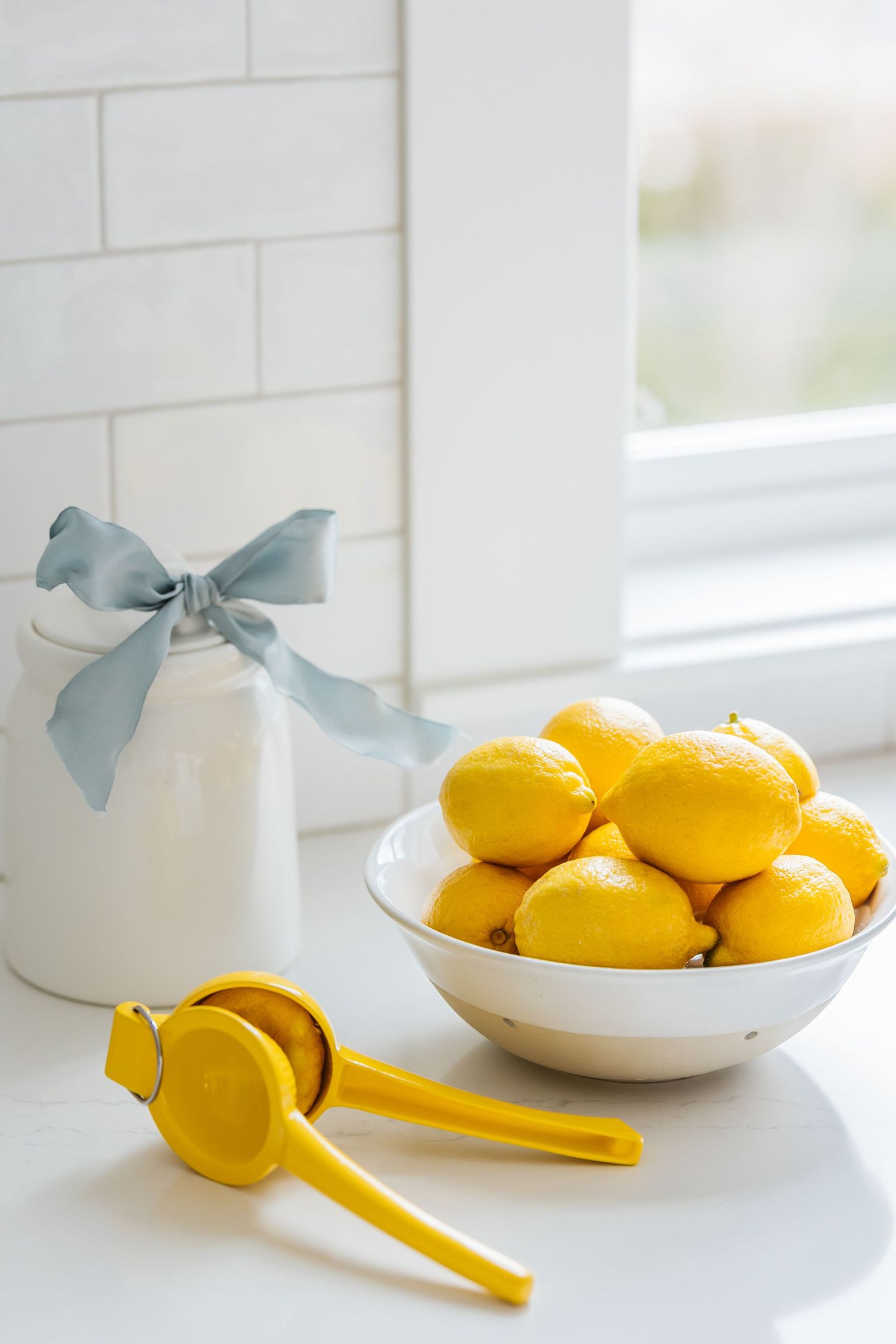 Bowl of fresh yellow lemons beside a citrus juicer and white ceramic jar with blue bow on a bright kitchen counter.