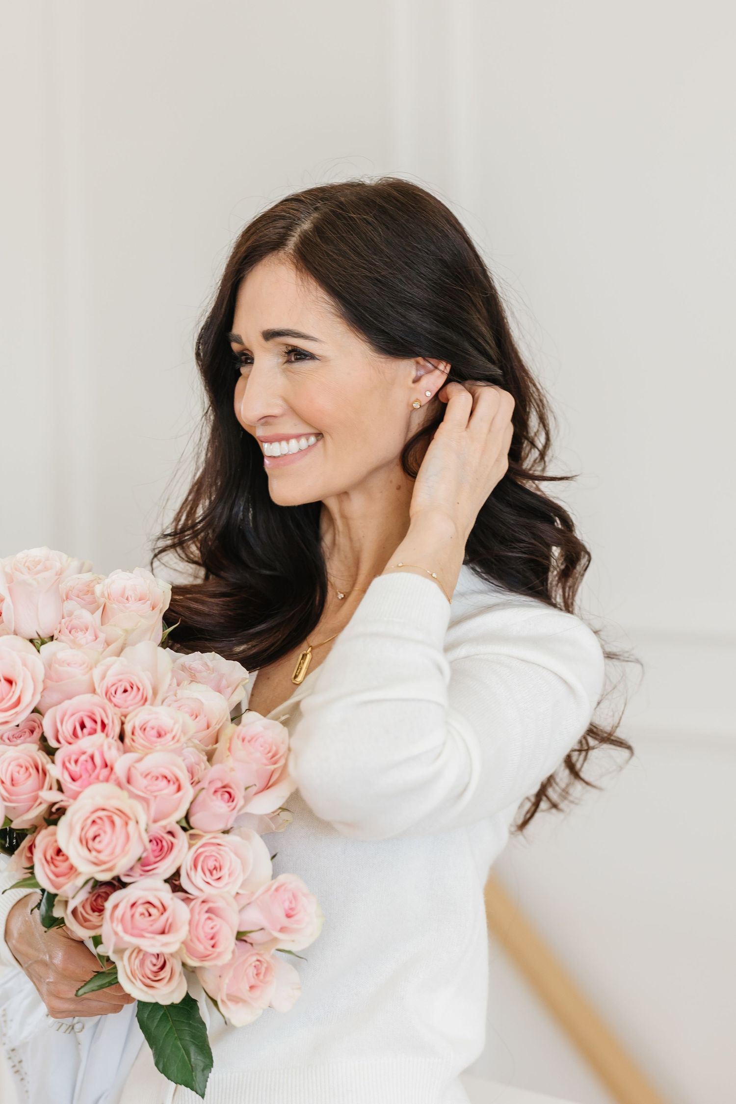 Smiling brunette woman in white top touching pearl earring while holding bouquet of pink roses.