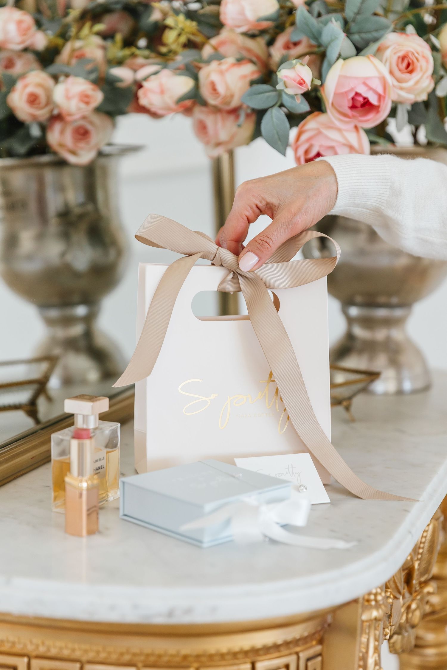 Hand holding a blush gift bag with gold ribbon, perfume, and jewelry box on marble table with pink roses.