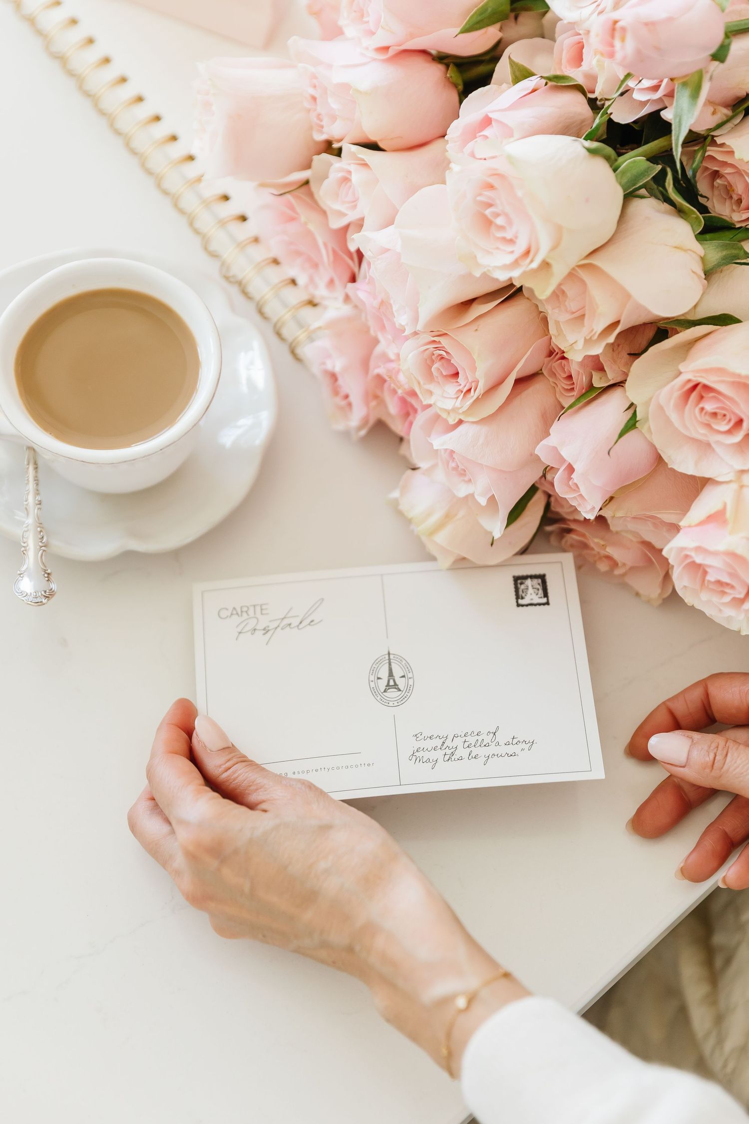 Hands holding a French postcard near pink roses bouquet and coffee cup on white desk.