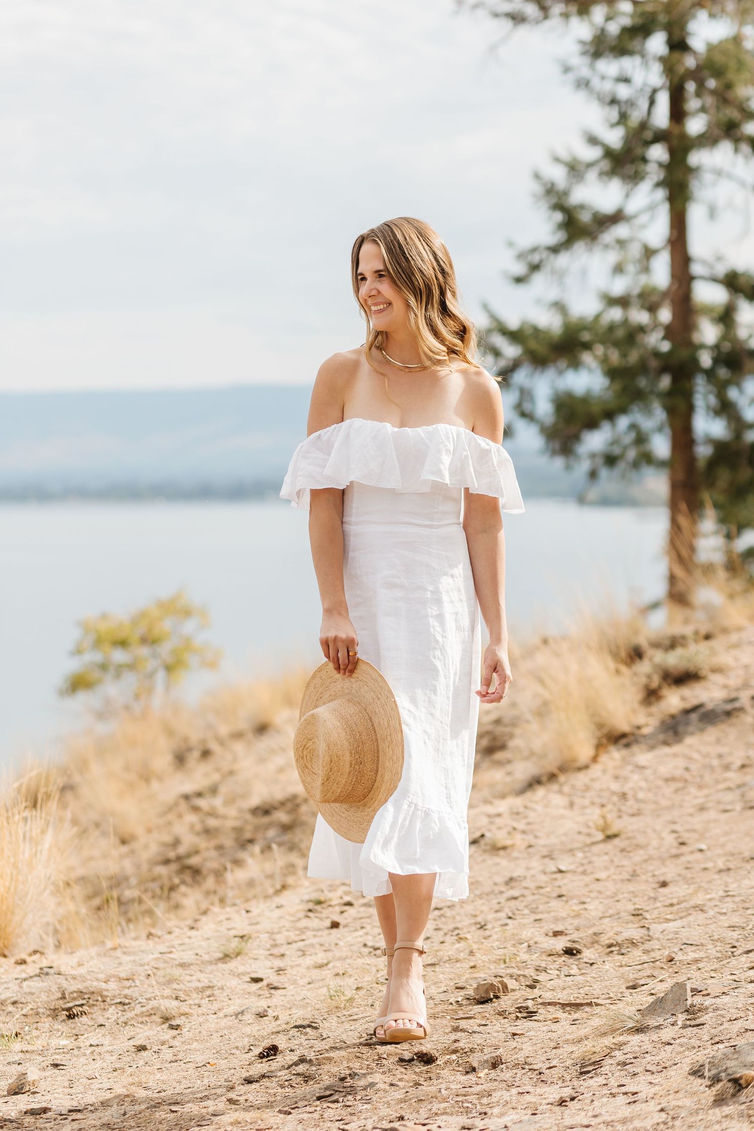 Woman in white summer dress walking along scenic mountain trail holding straw hat.