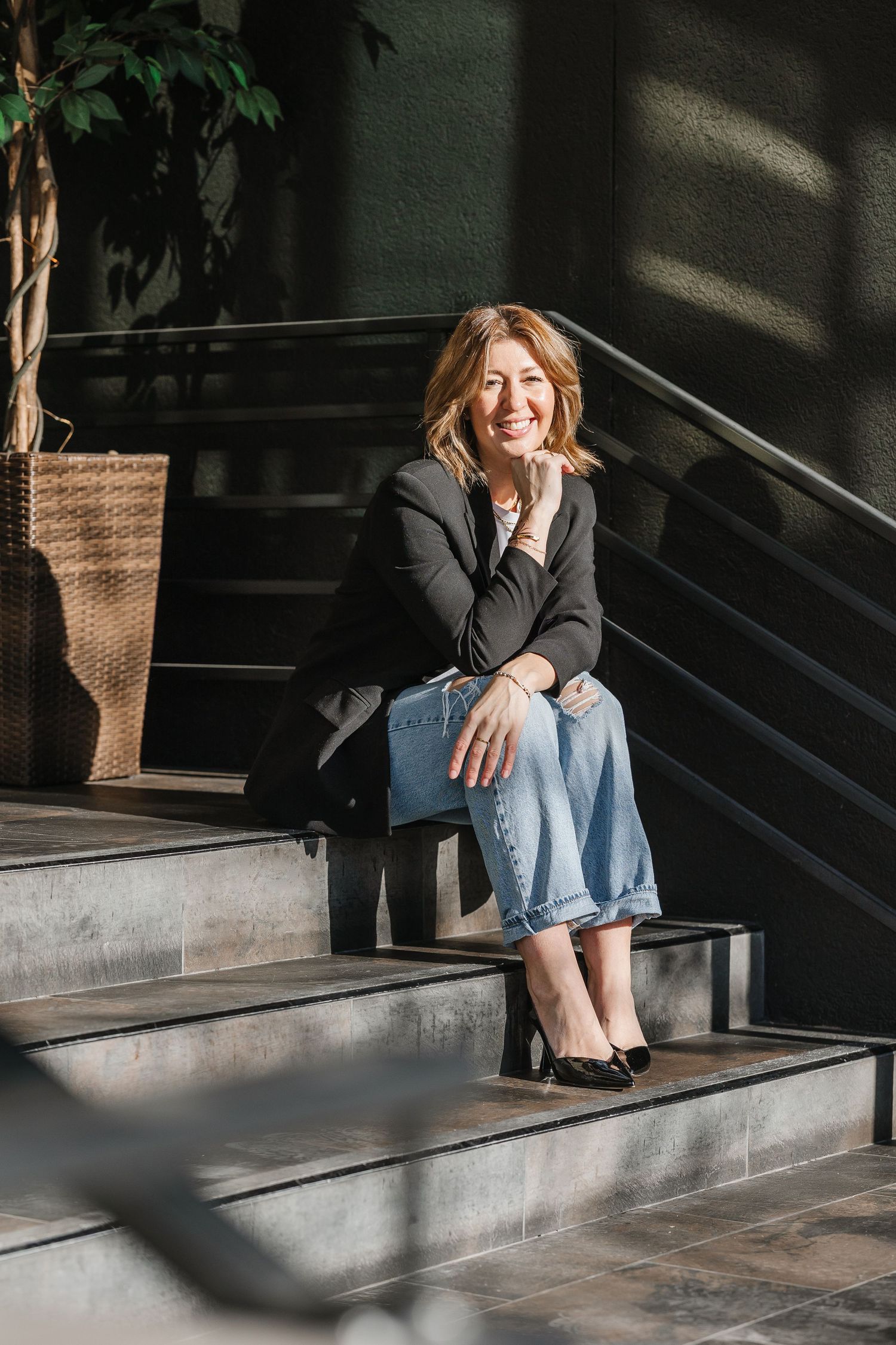 Woman in black blazer and jeans smiling while sitting on modern steps beside a potted plant.