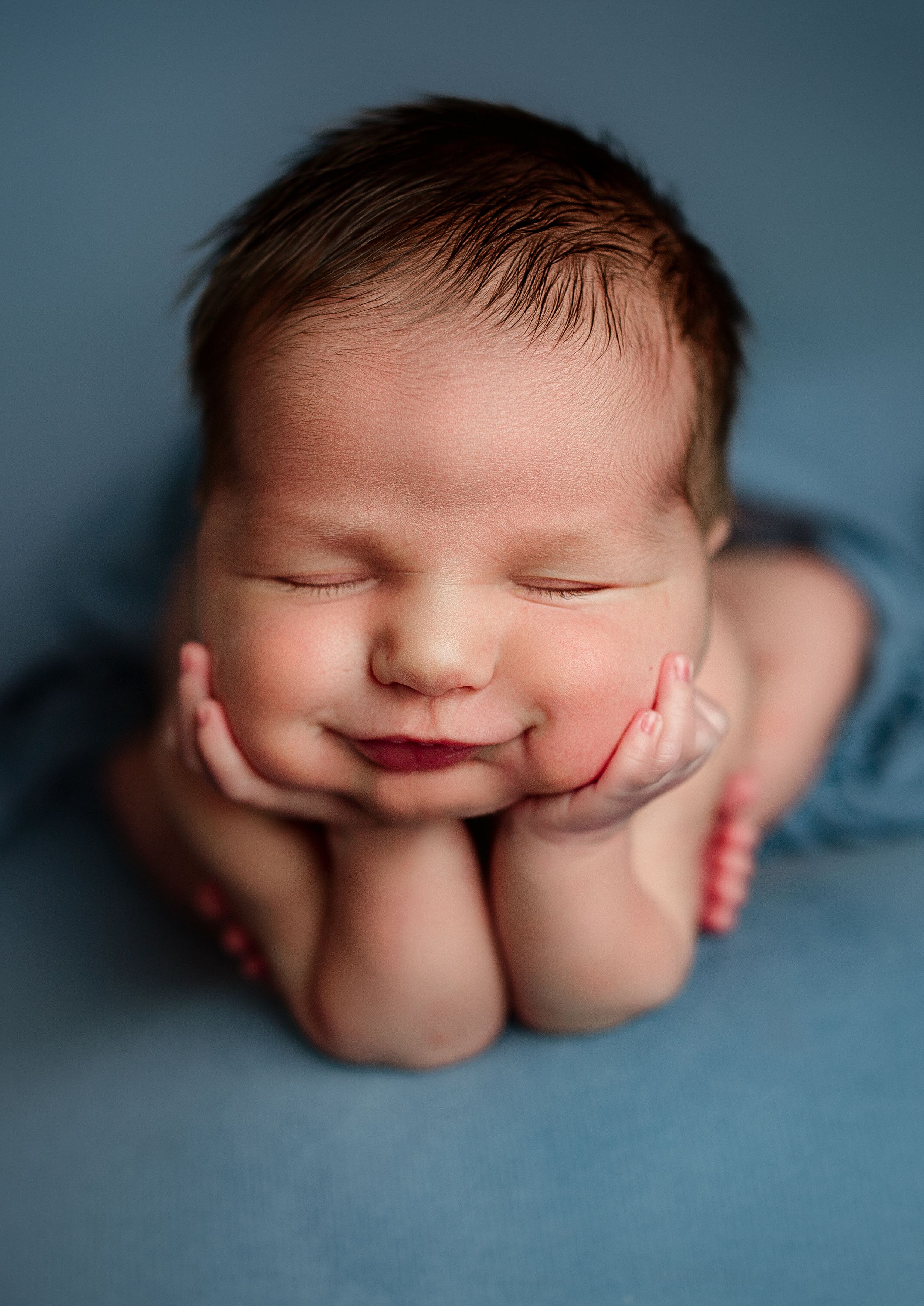 Peaceful sleeping newborn baby posed with chin resting on hands in classic froggy position on blue backdrop.