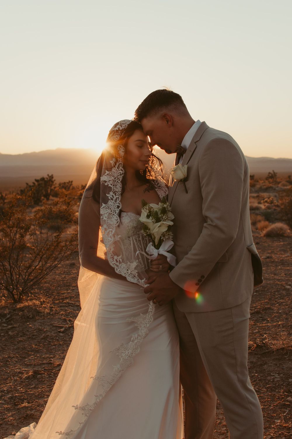 A romantic desert wedding photoshoot at sunset with soft golden light illuminating the couple in an intimate embrace.