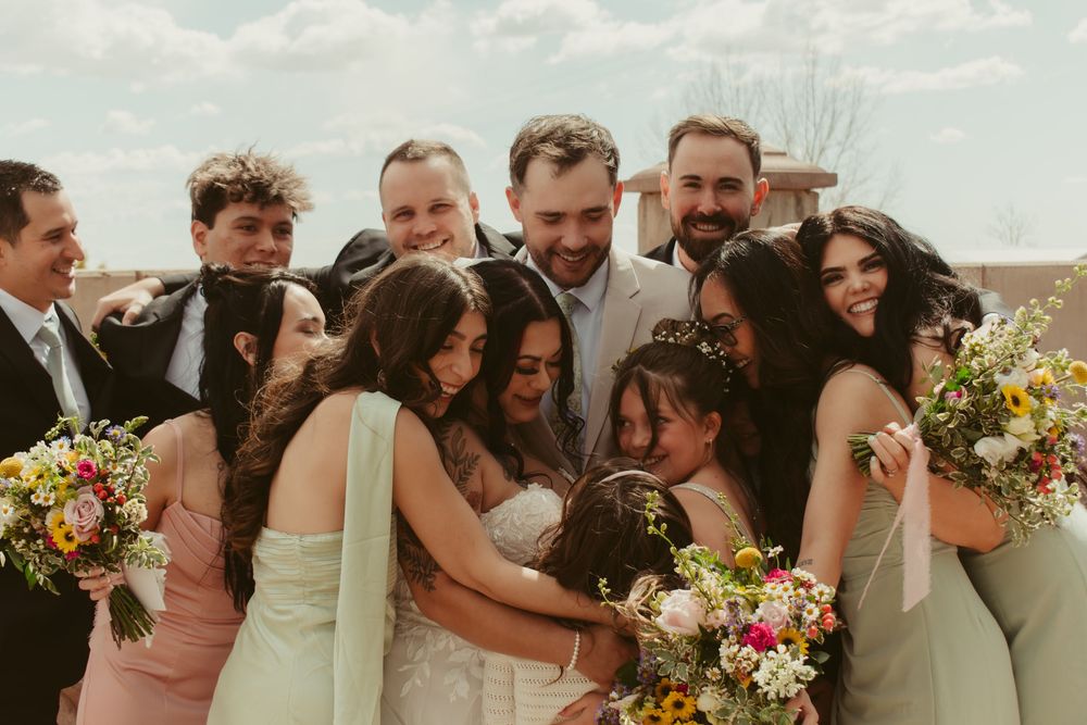 Wedding party laughs together while standing in a line outdoors with colorful bouquets and formal attire.