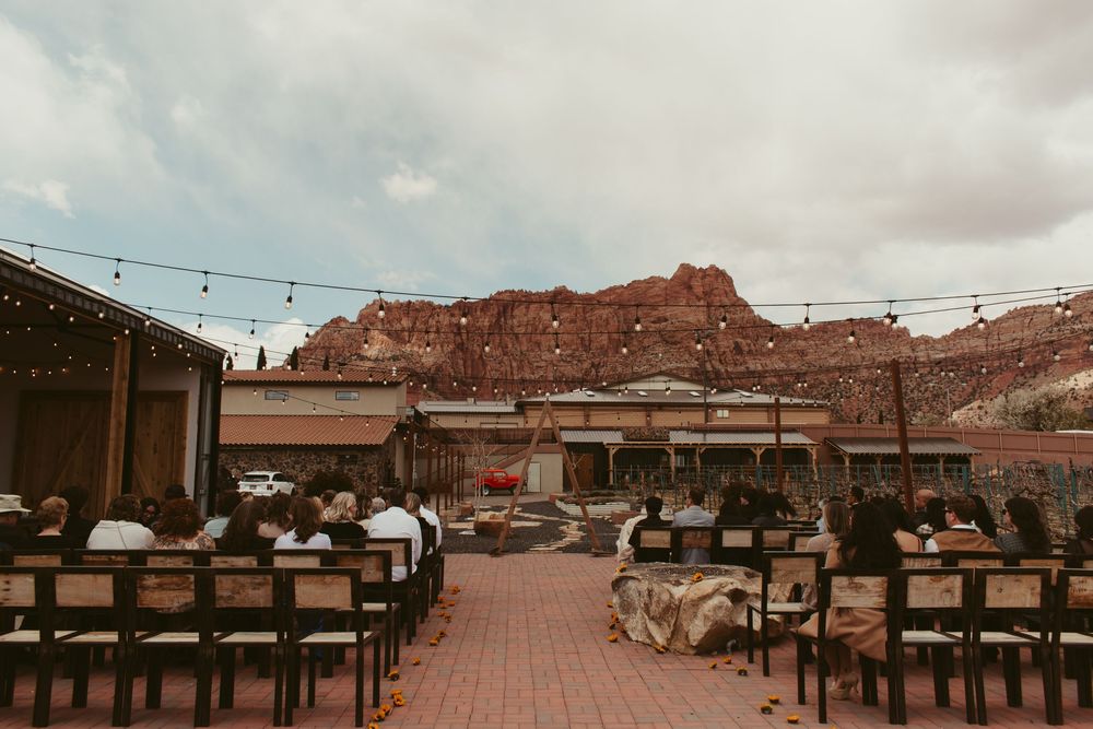 Outdoor wedding venue setup with wooden chairs and string lights against dramatic red rock formations in the desert landscape.
