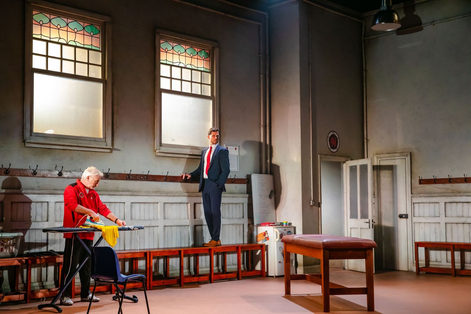Two performers interact in sparse room setting with vintage windows and wooden chairs.