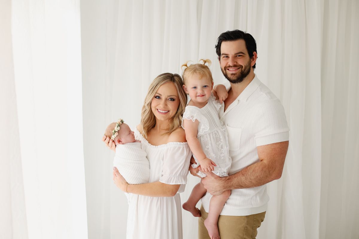 Family wearing all white smiling at the camera, mom holding baby, dad holding toddler girl