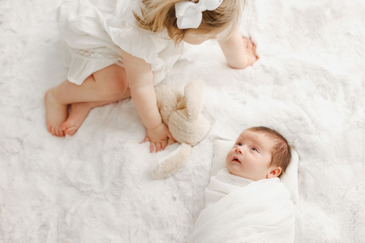 toddler sister holding a toy bunny next to her newborn brother on a white fur blanket