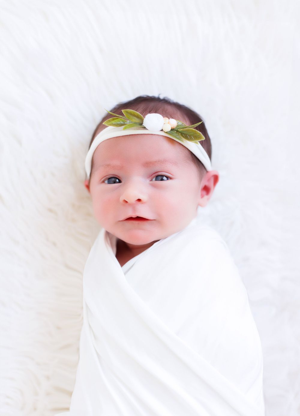 Newborn baby wrapped in white blanket wearing floral headband on white background.