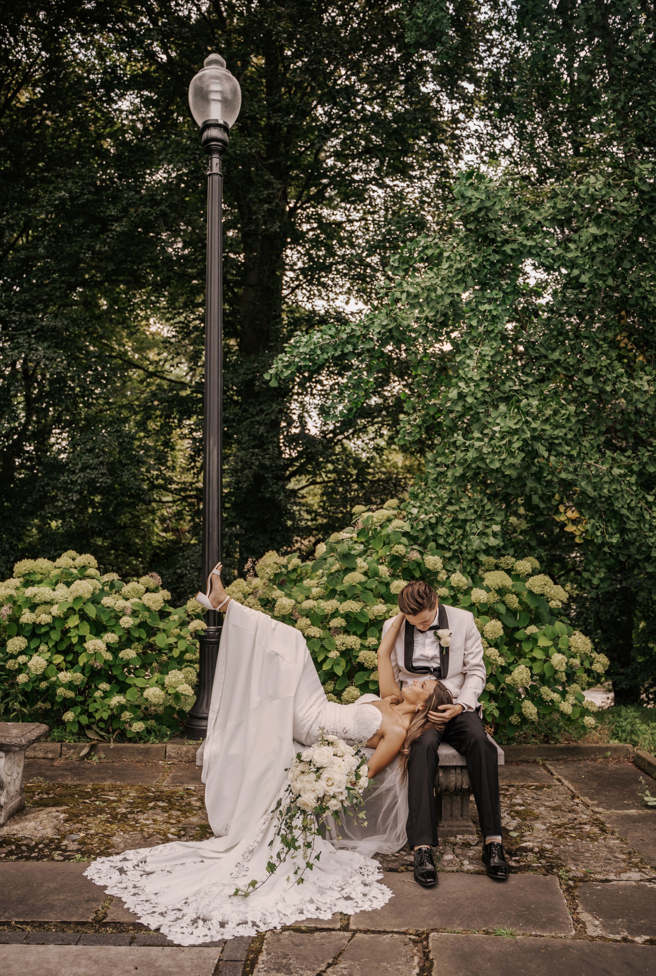 A couple shares a romantic moment on stone steps surrounded by lush greenery and vintage lampposts in an outdoor garden setting.