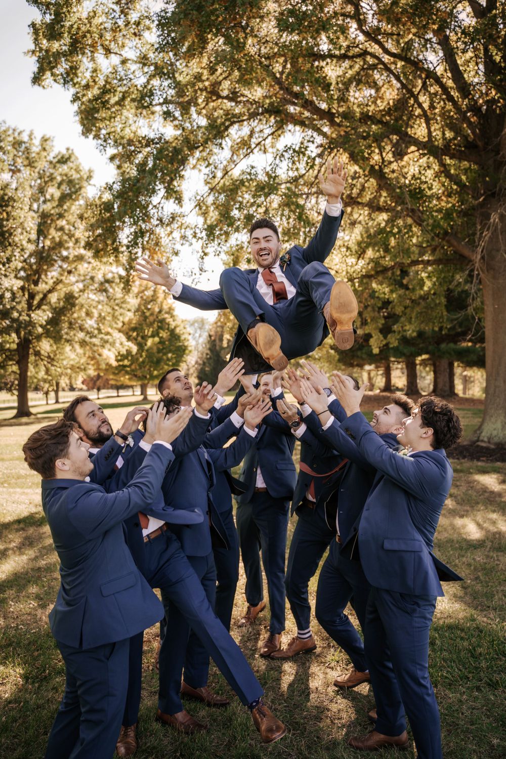 Groomsmen in navy suits tossing groom in air during outdoor wedding photo.