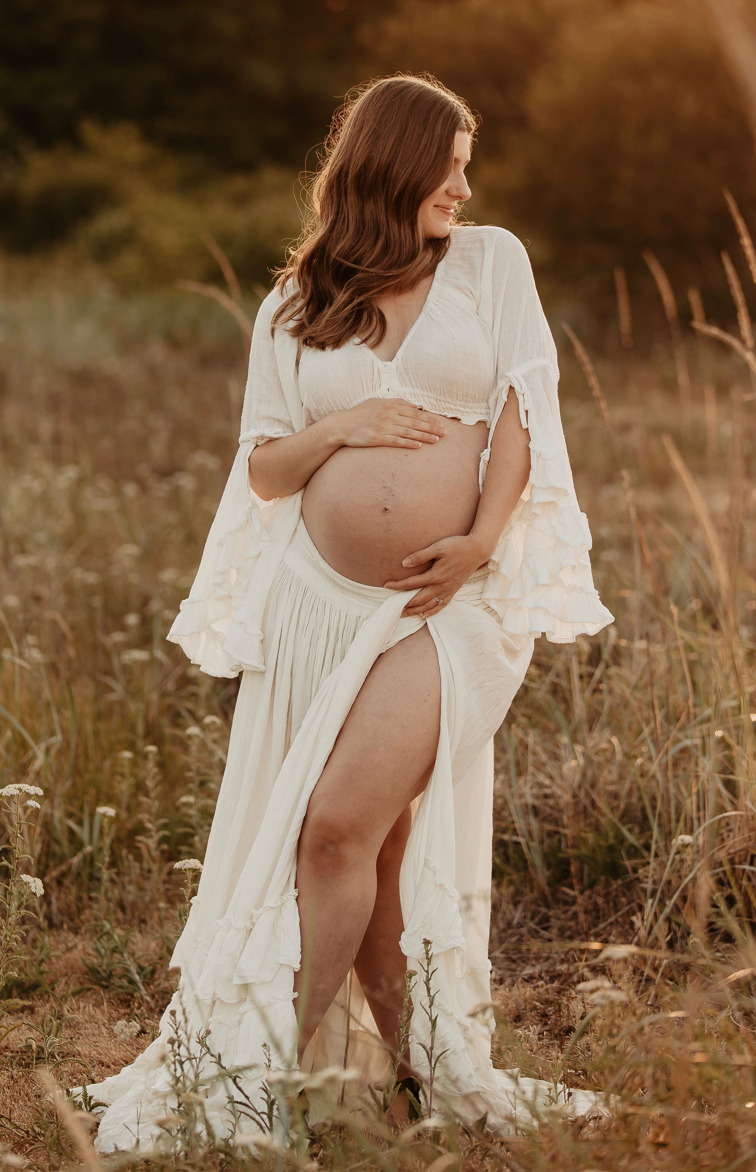 Pregnant woman in white boho maternity gown posing in a sunlit field, cradling her baby bump.