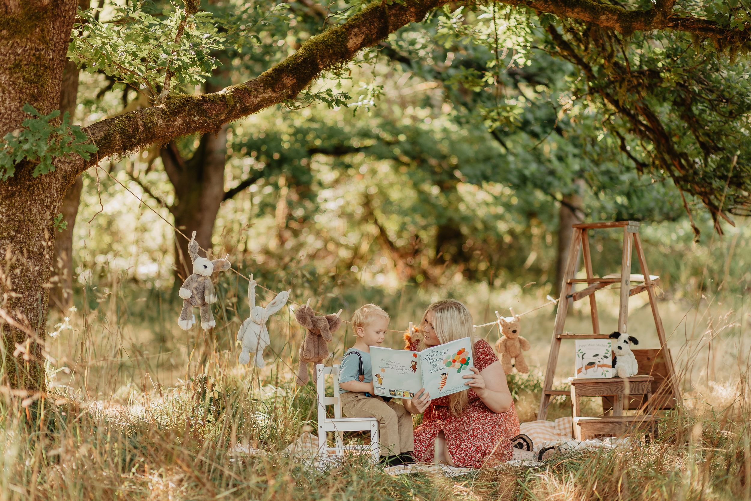 Two children enjoy a whimsical outdoor tea party under a large tree surrounded by stuffed animals in a golden meadow.