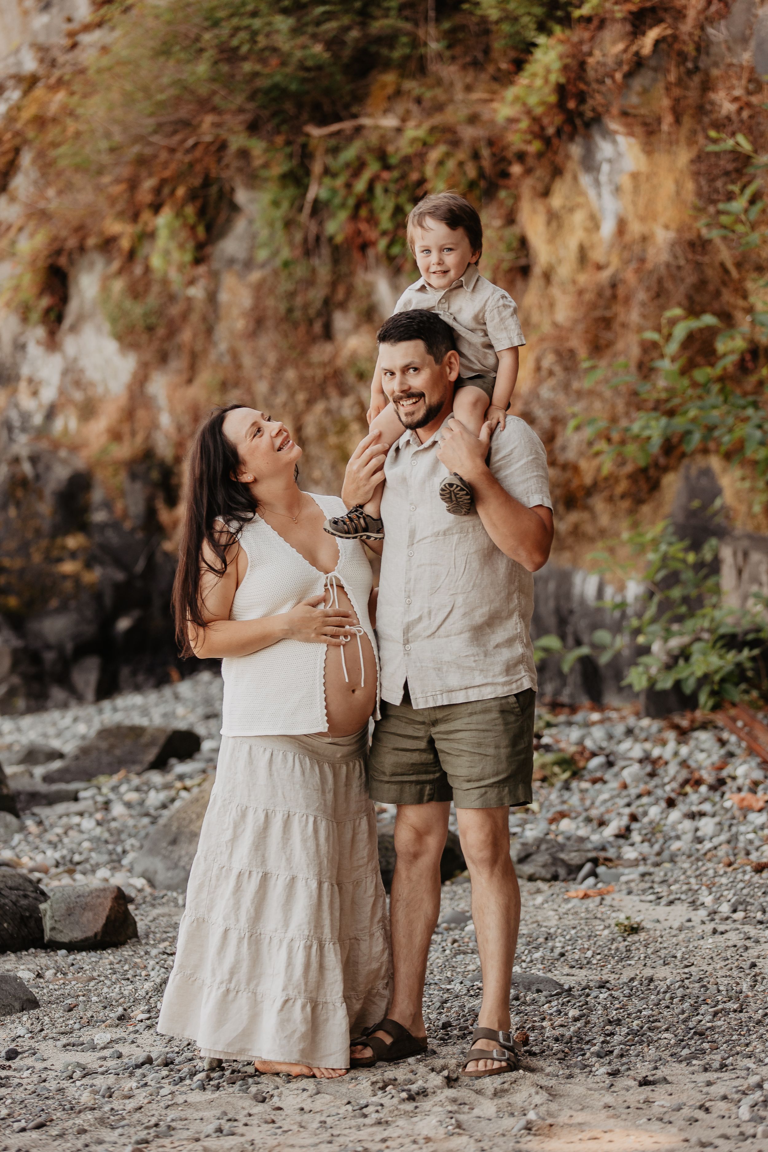 Pregnant woman in white dress with husband and toddler son on rocky beach, warm family maternity photo.