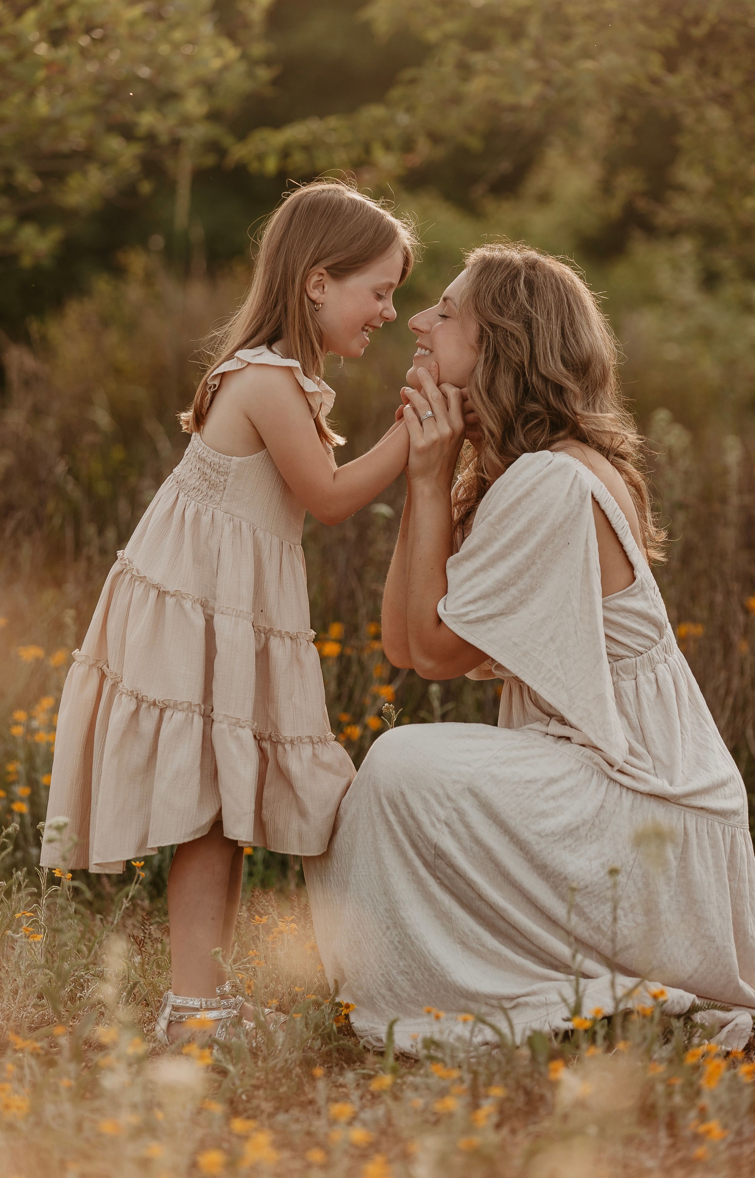 A mother and young daughter share a joyful moment in a sunny wildflower field, both wearing flowing beige dresses.