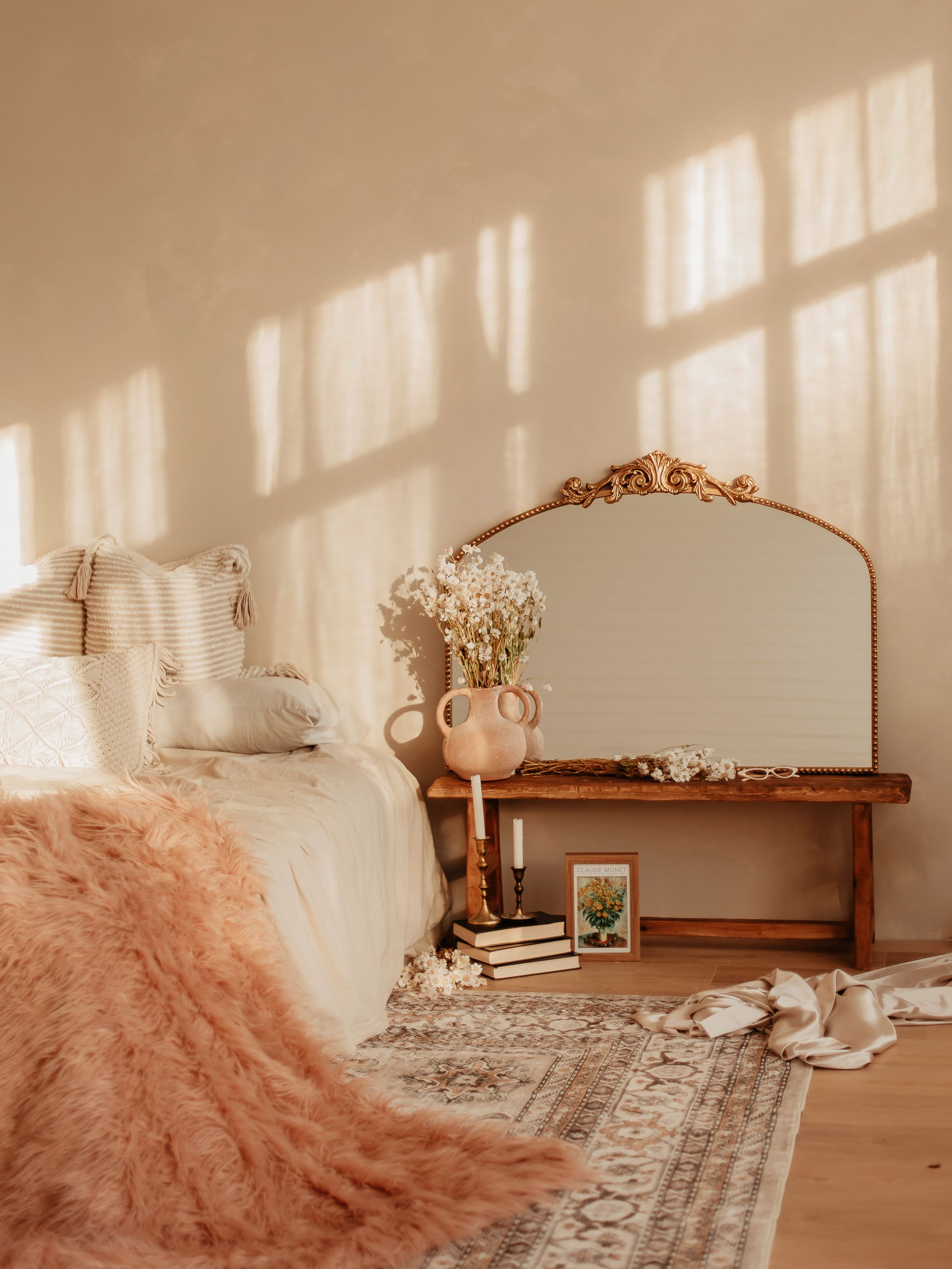 Boho bedroom with ornate gold mirror, wooden bench, pink throw blanket, vintage rug, and warm sunlight casting window shadows.